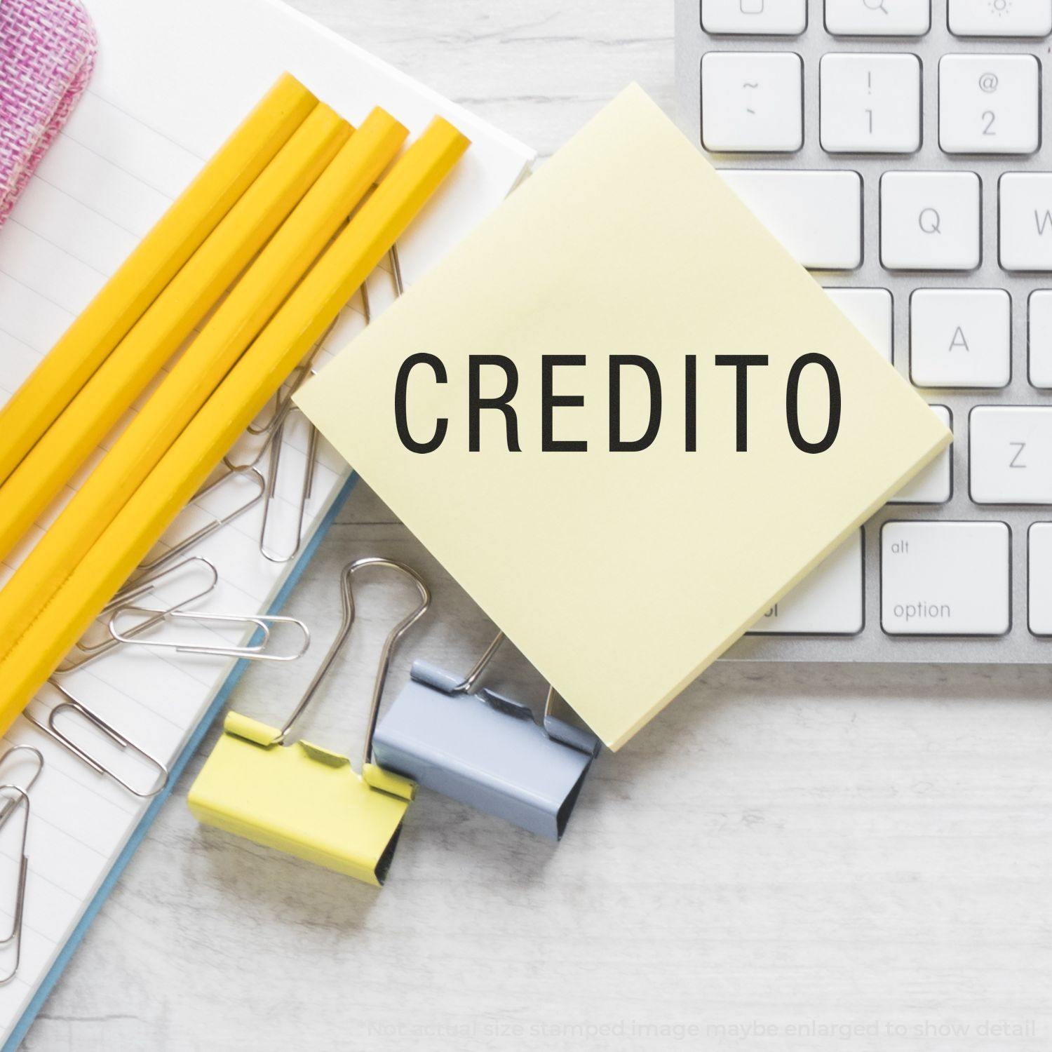 Slim Pre-Inked Credito Stamp on a yellow sticky note beside pencils, paper clips, and a keyboard on a desk.