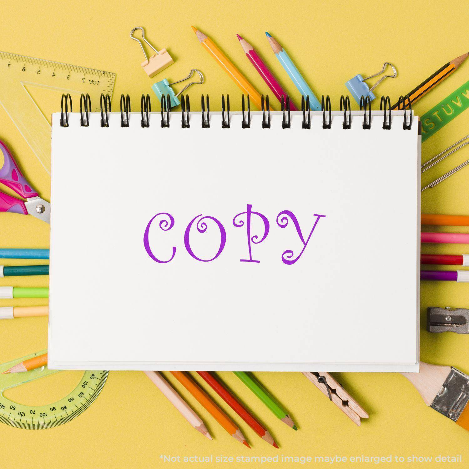 Large Pre-Inked Curly Copy Stamp imprint on a white notebook surrounded by colorful pencils, rulers, and other stationery on a yellow background.
