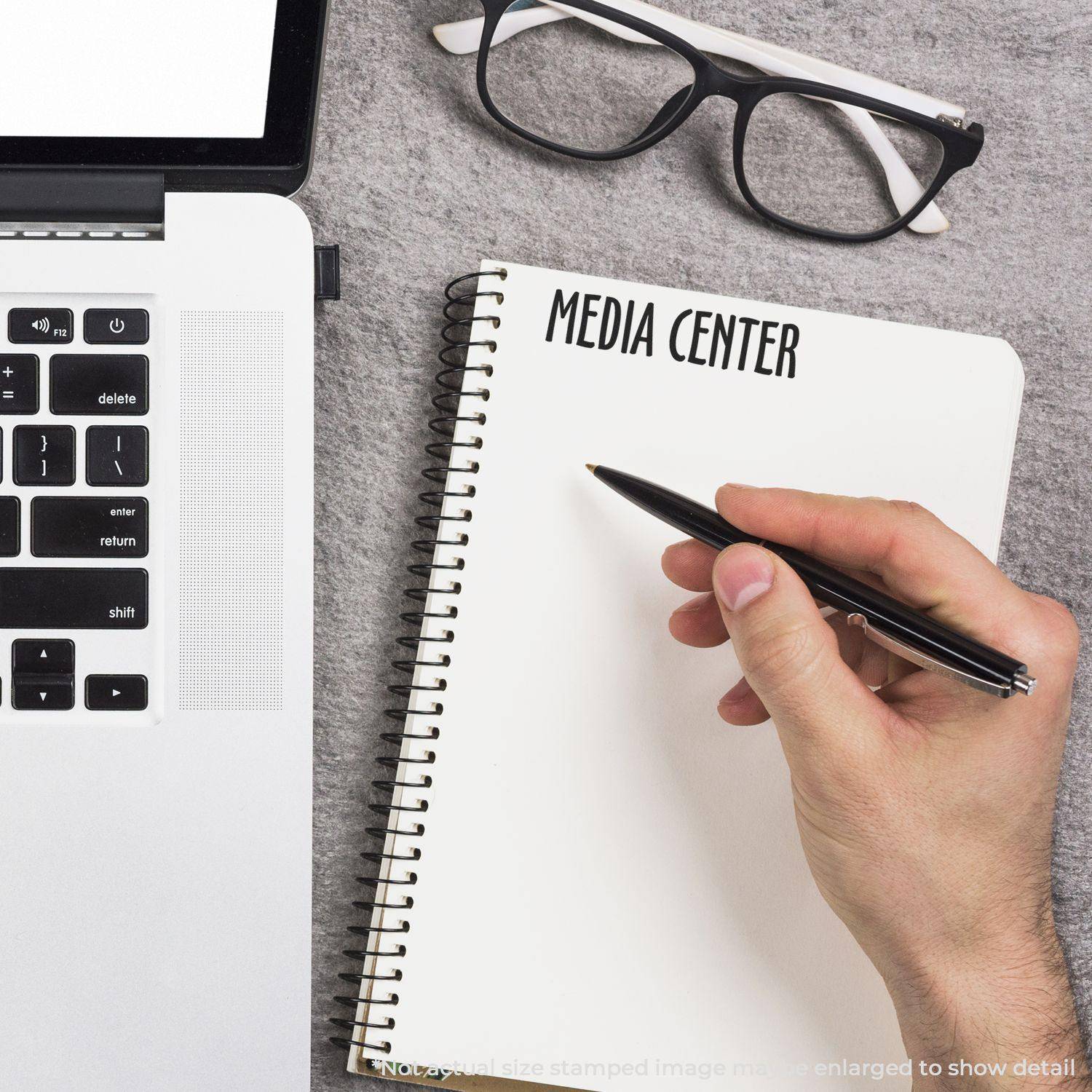 Hand holding pen near notebook with MEDIA CENTER stamped using Large Pre-Inked Media Center Stamp, next to a laptop and glasses.