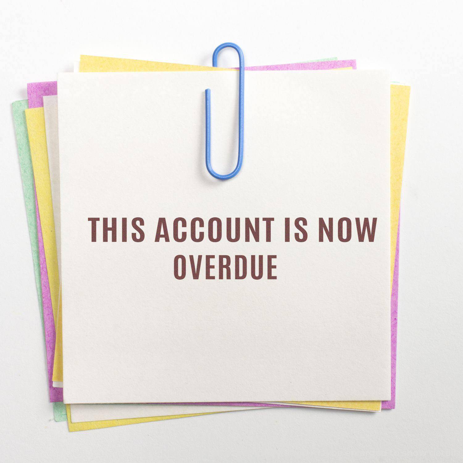 Large Pre-Inked Narrow This Account Is Now Overdue Stamp on a stack of colorful papers clipped together with a blue paperclip.
