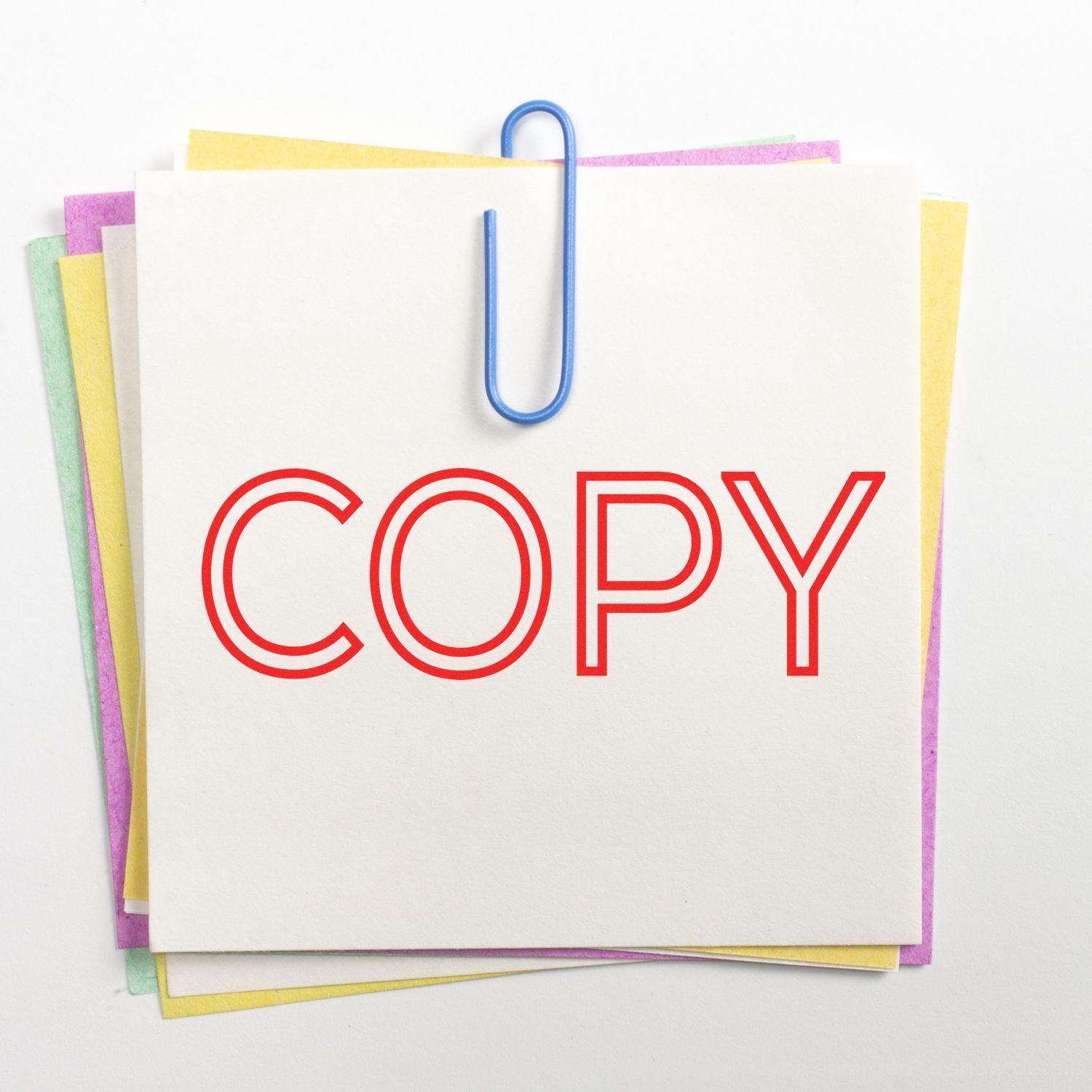 Outline Copy Rubber Stamp in red ink on a white paper clipped with a blue paperclip, stacked on colored papers.