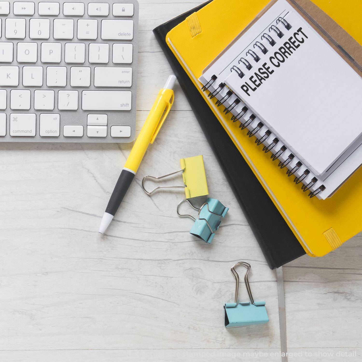 A desk with a keyboard, pen, binder clips, and a notebook stamped with PLEASE CORRECT using the Large Please Correct Rubber Stamp.