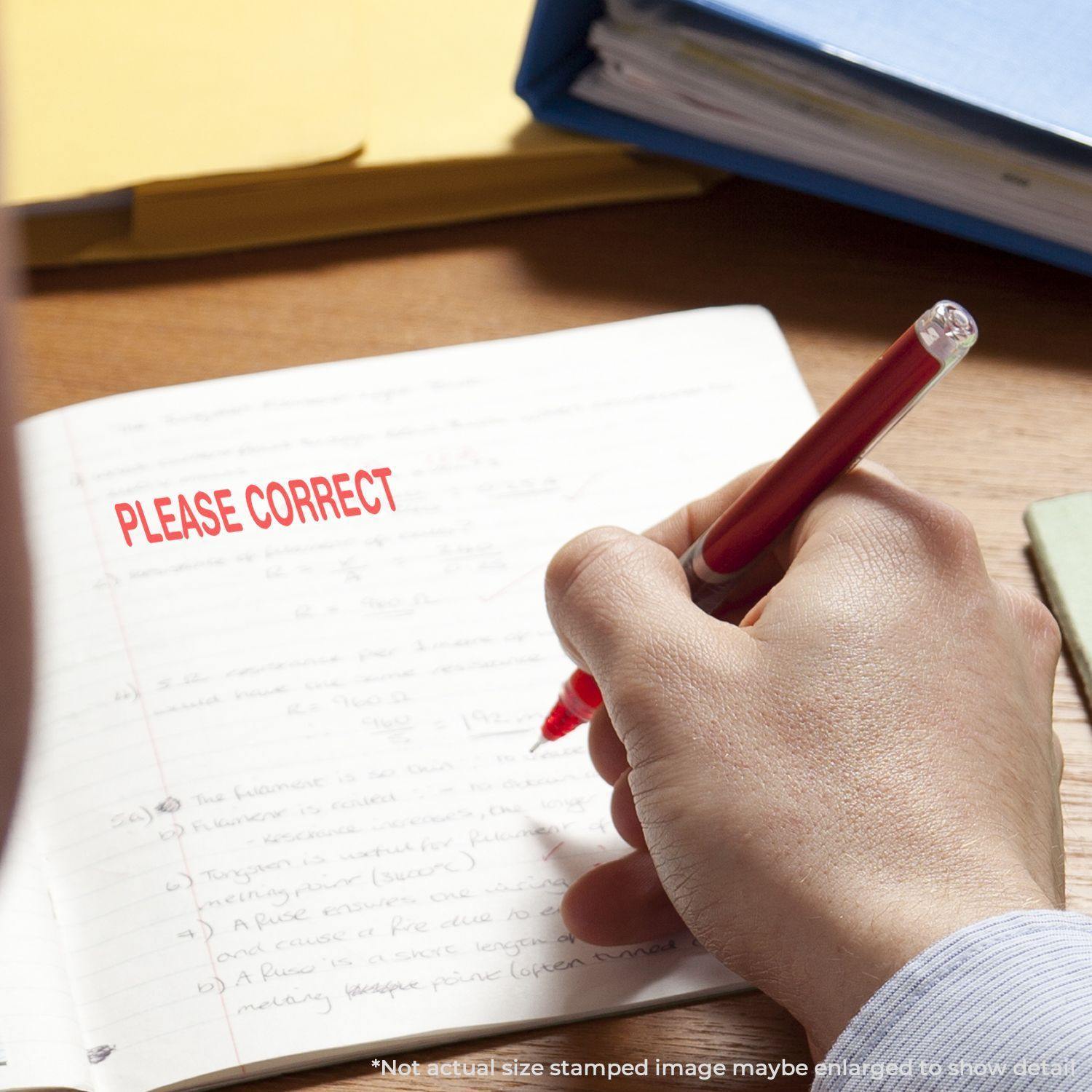A hand holding a red pen next to a Slim Pre-Inked Please Correct Teacher Stamp on a notebook with PLEASE CORRECT stamped in red.