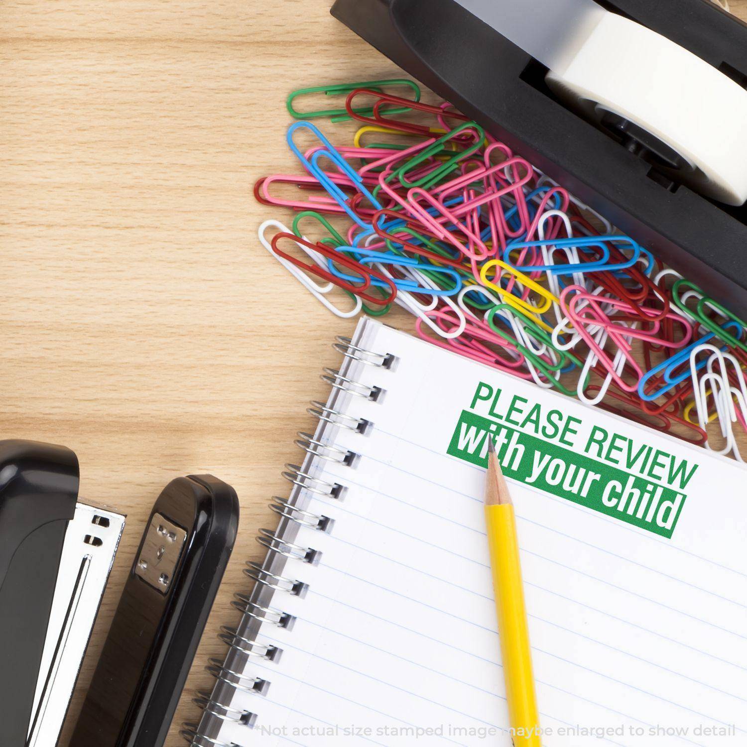 A desk with a stapler, tape dispenser, colorful paperclips, and a notebook stamped with Large Please Review with your child Rubber Stamp in green.