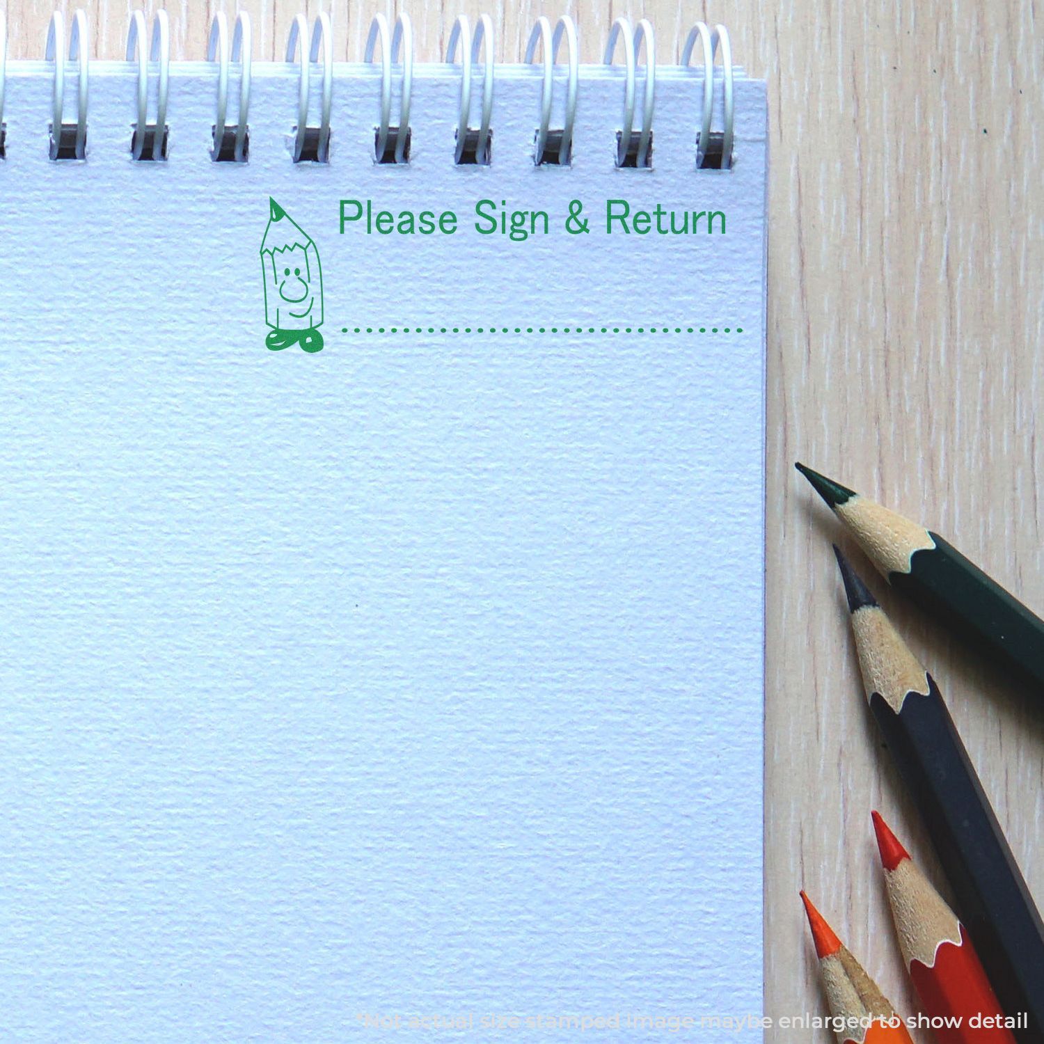 Self Inking Please Sign and Return Stamp in green ink on a spiral notebook, with colored pencils beside it on a wooden surface.