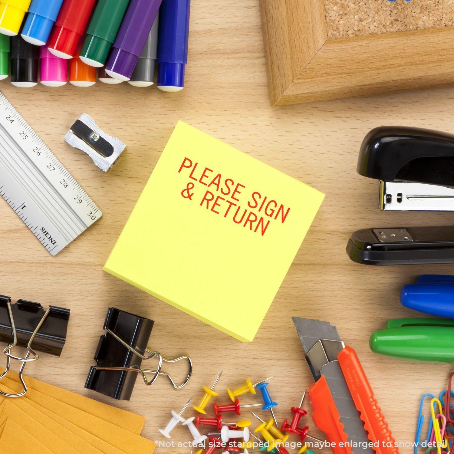 Please Sign & Return rubber stamp on a yellow sticky note surrounded by office supplies like pens, stapler, and push pins.