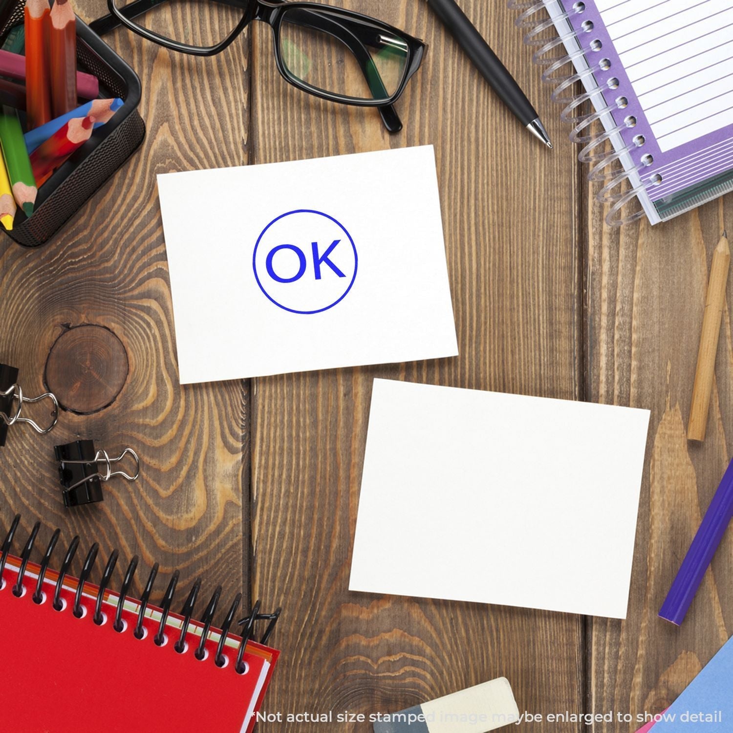 A Self Inking Round OK Stamp on a white card, surrounded by office supplies including notebooks, glasses, and pens on a wooden desk.