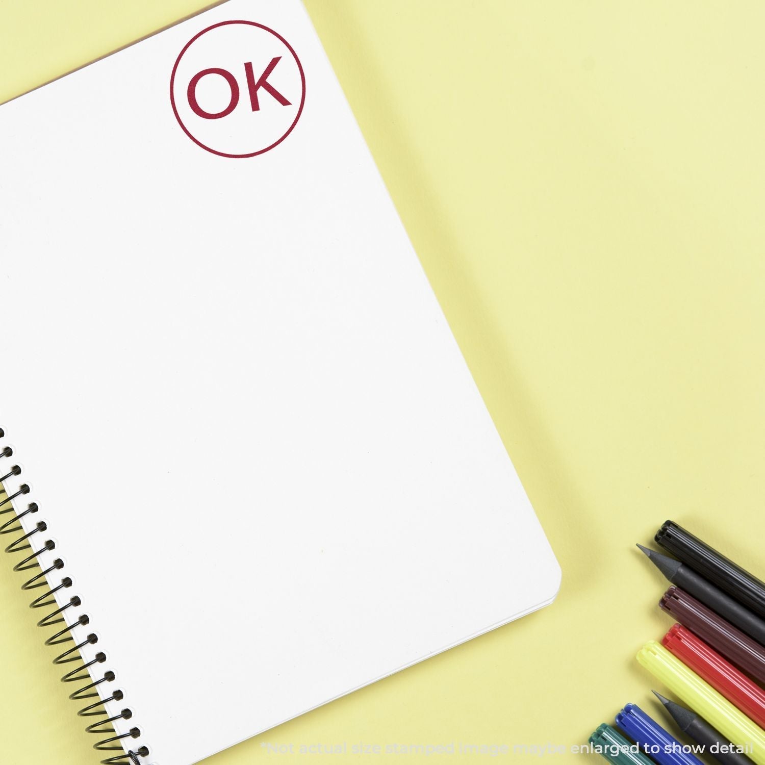 Self Inking Round OK Stamp marking OK on a white spiral notebook, with colorful pens on a yellow background.