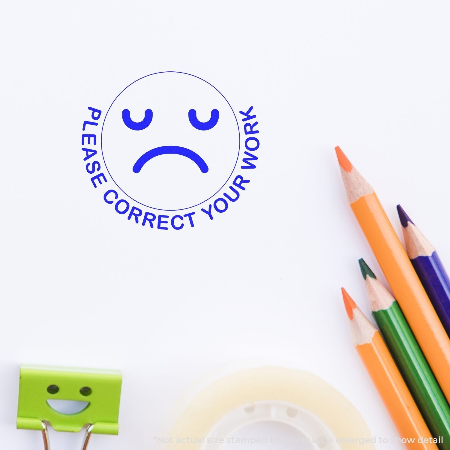 Self Inking Round Please Correct Smiley Stamp with a sad face imprint on paper, surrounded by colorful pencils, tape, and a smiley clip.