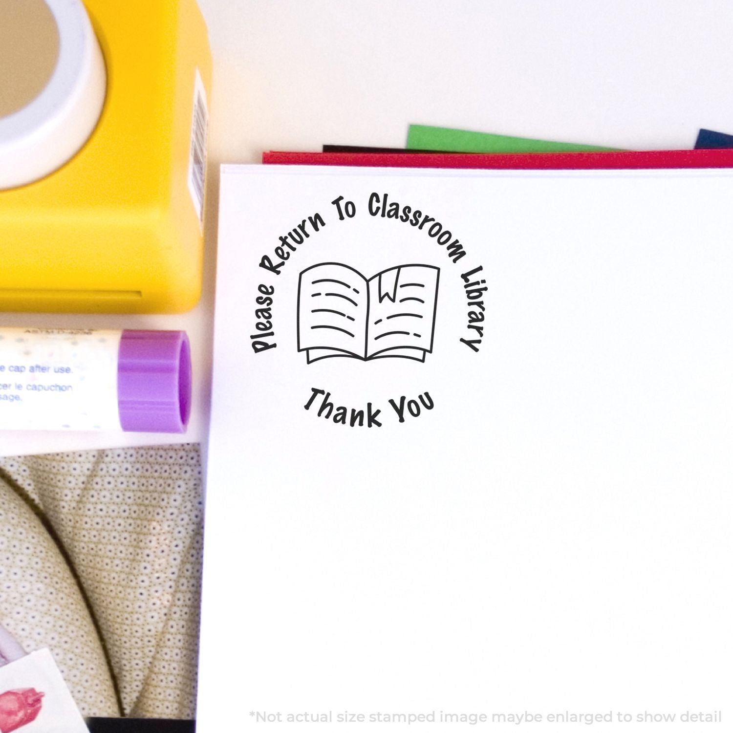 Self Inking Round Please Return to Classroom Library Stamp on a white paper, surrounded by a yellow box, purple glue stick, and other stationery.
