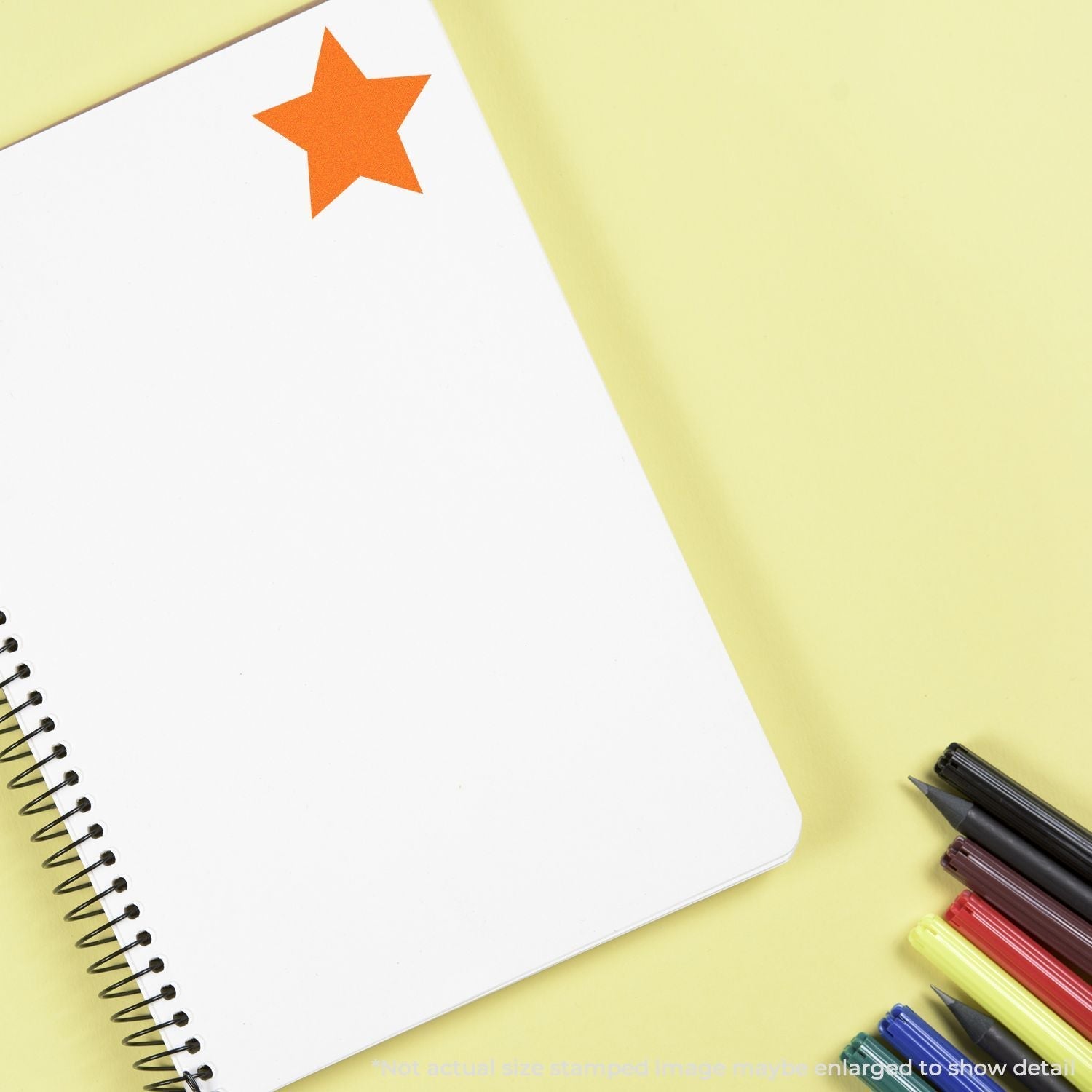 Self Inking Round Solid Star Stamp in orange used on a white spiral notebook, with colored pens placed nearby on a yellow background.