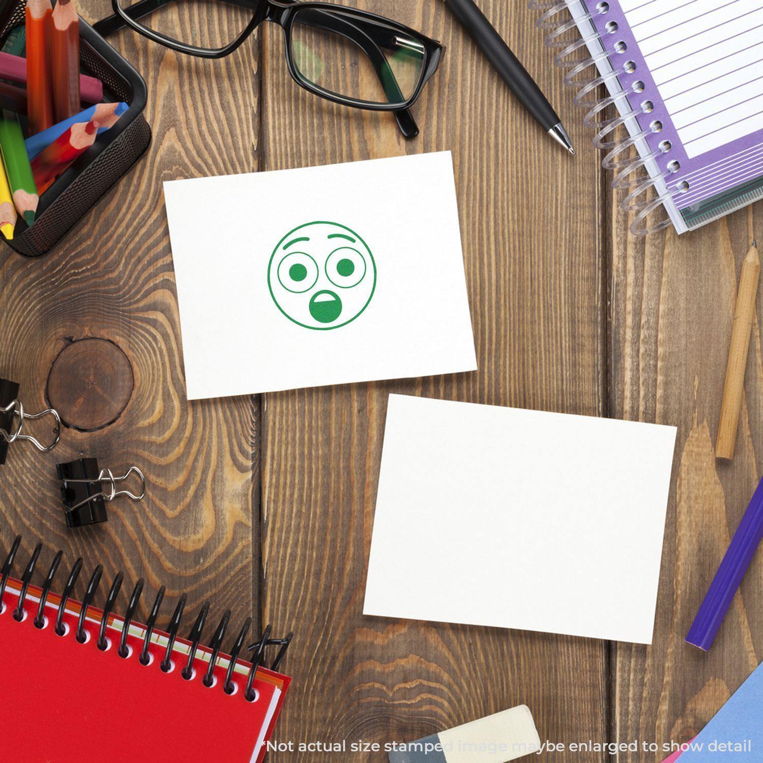 Round Surprised Smiley Rubber Stamp on white paper, surrounded by office supplies on a wooden desk.