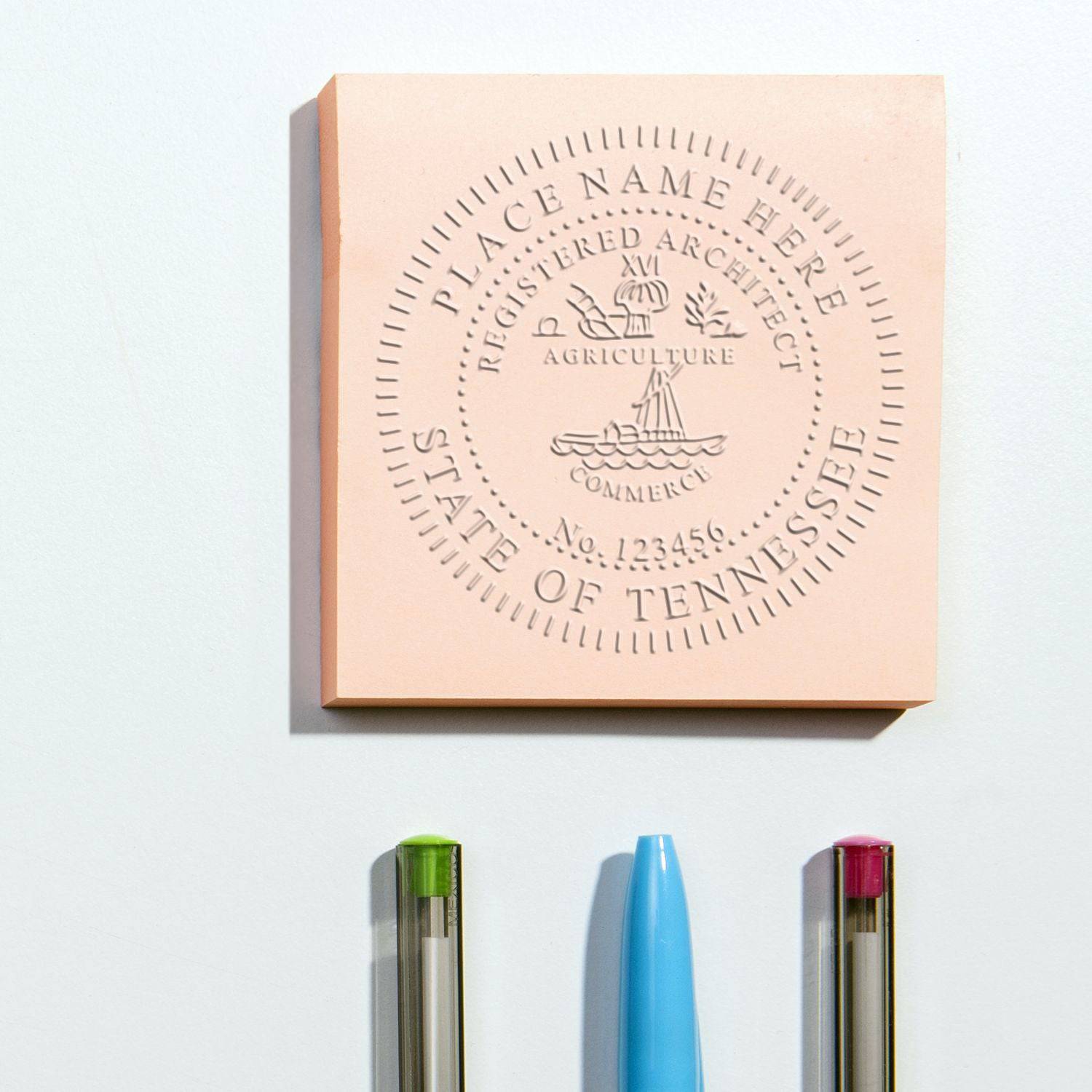 Architect Handheld Seal Embosser imprint on a pink square paper, displaying State of Tennessee and Registered Architect with a seal design. Three pens are placed below the embossed paper.