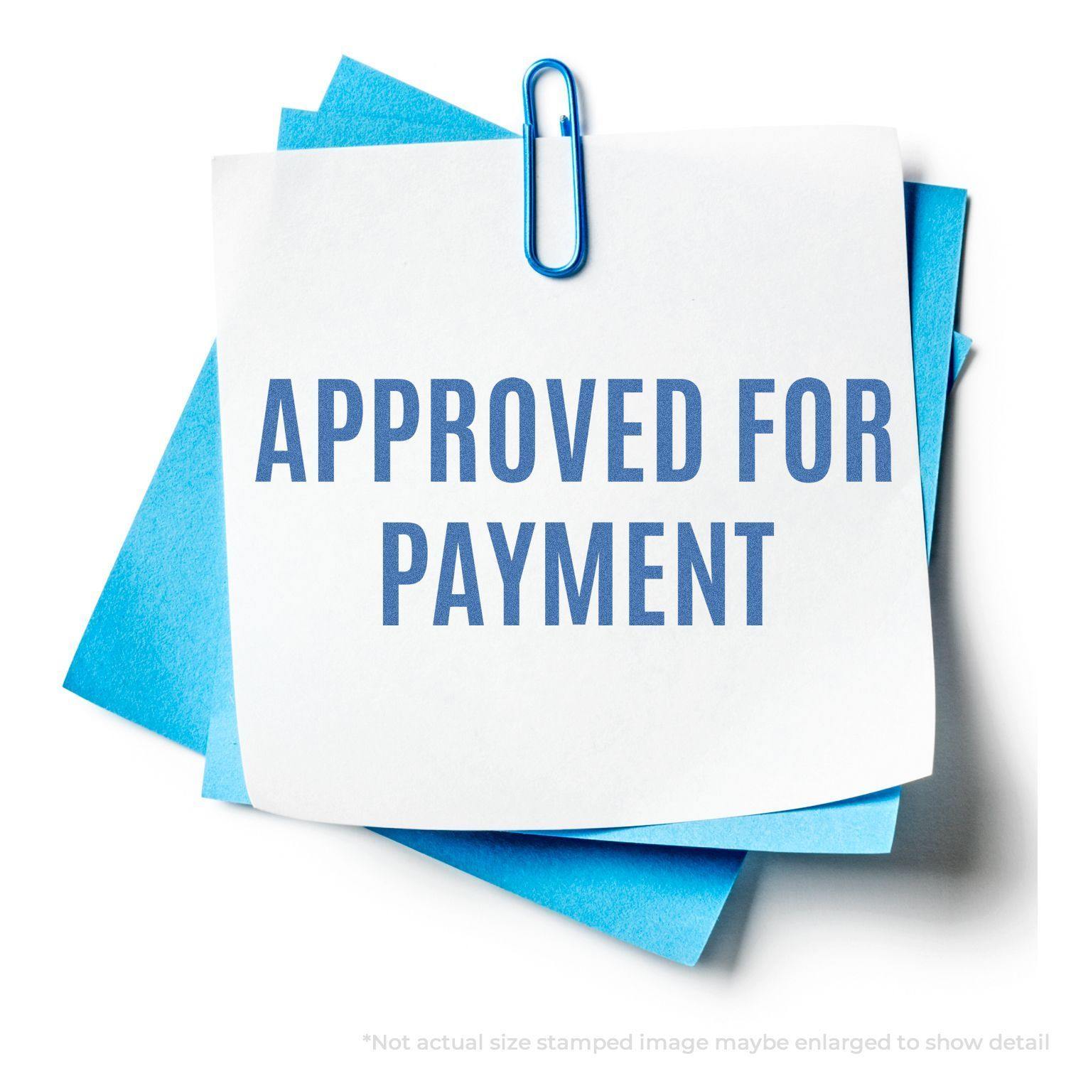 A blue paperclip holds a white note with Approved For Payment stamped on it using the Approved For Payment Xstamper Stamp, over blue papers.