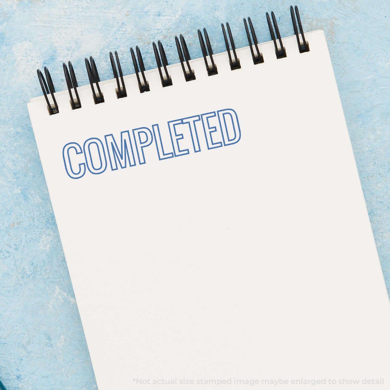 A Blue Completed Xstamper Stamp imprint on a white spiral notebook, showing the word COMPLETED in blue ink on a light blue background.