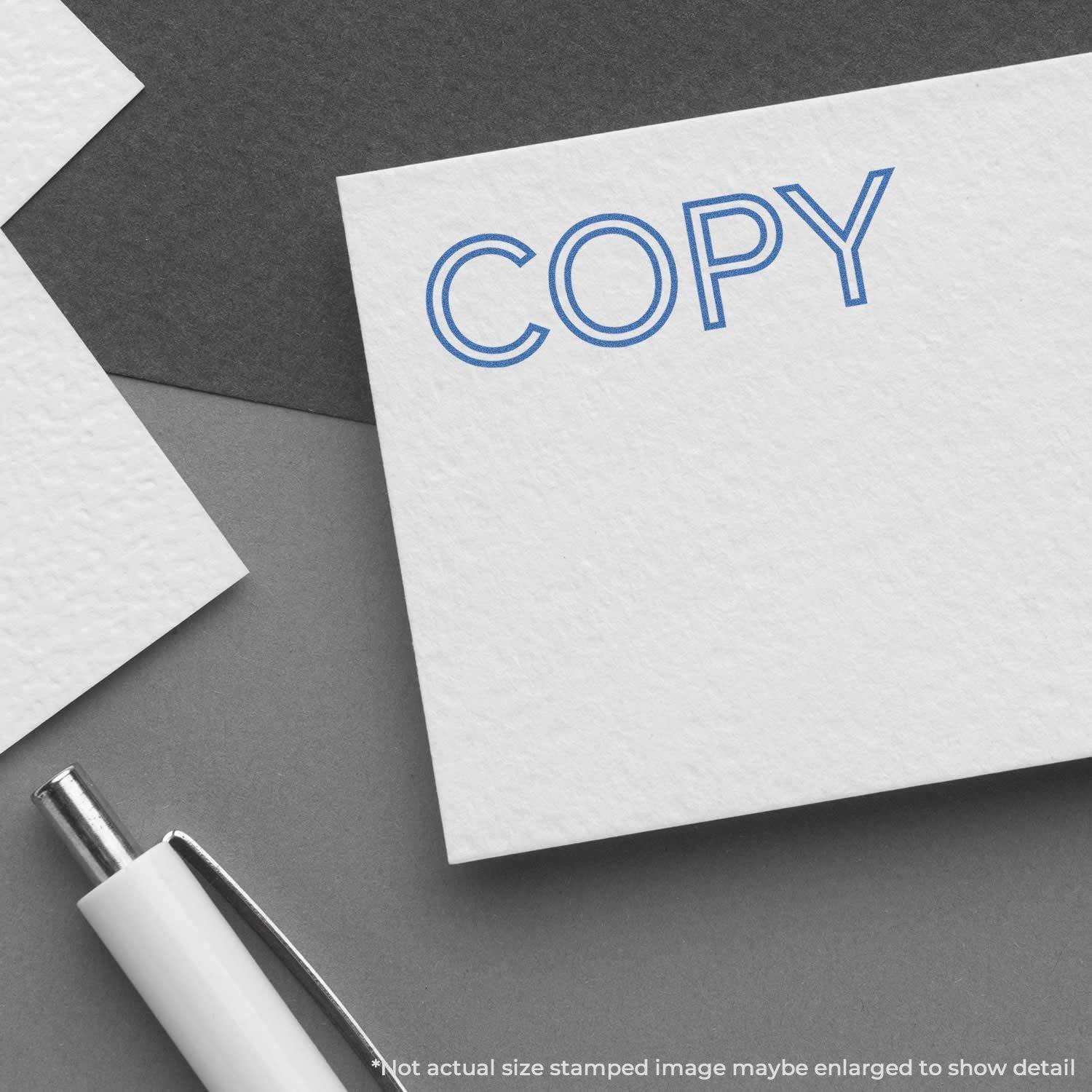 A Blue Copy Xstamper Stamp imprint on a white card, placed on a dark surface with a white pen nearby.
