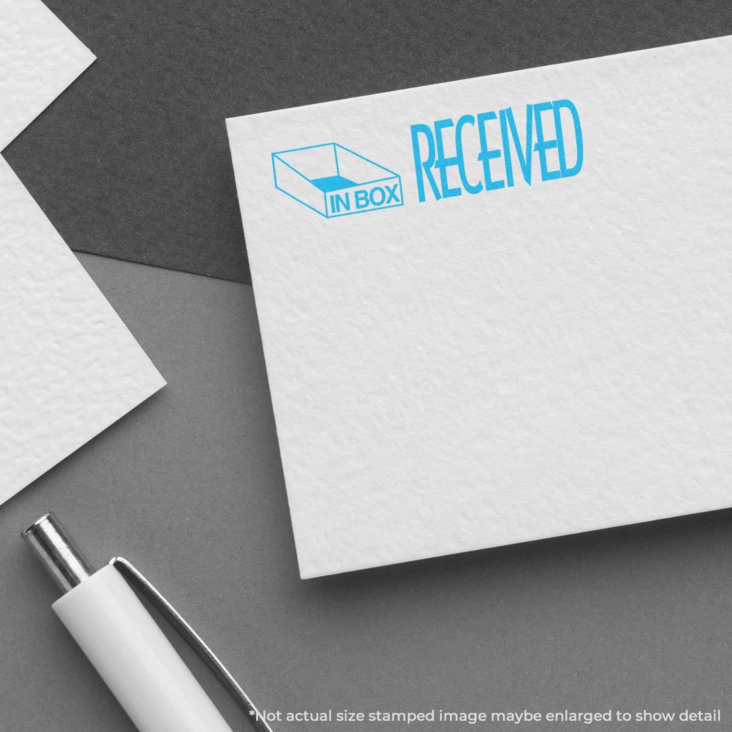 A white card stamped with IN BOX RECEIVED in blue using the Blue Received Xstamper Stamp, placed on a gray surface with a pen nearby.