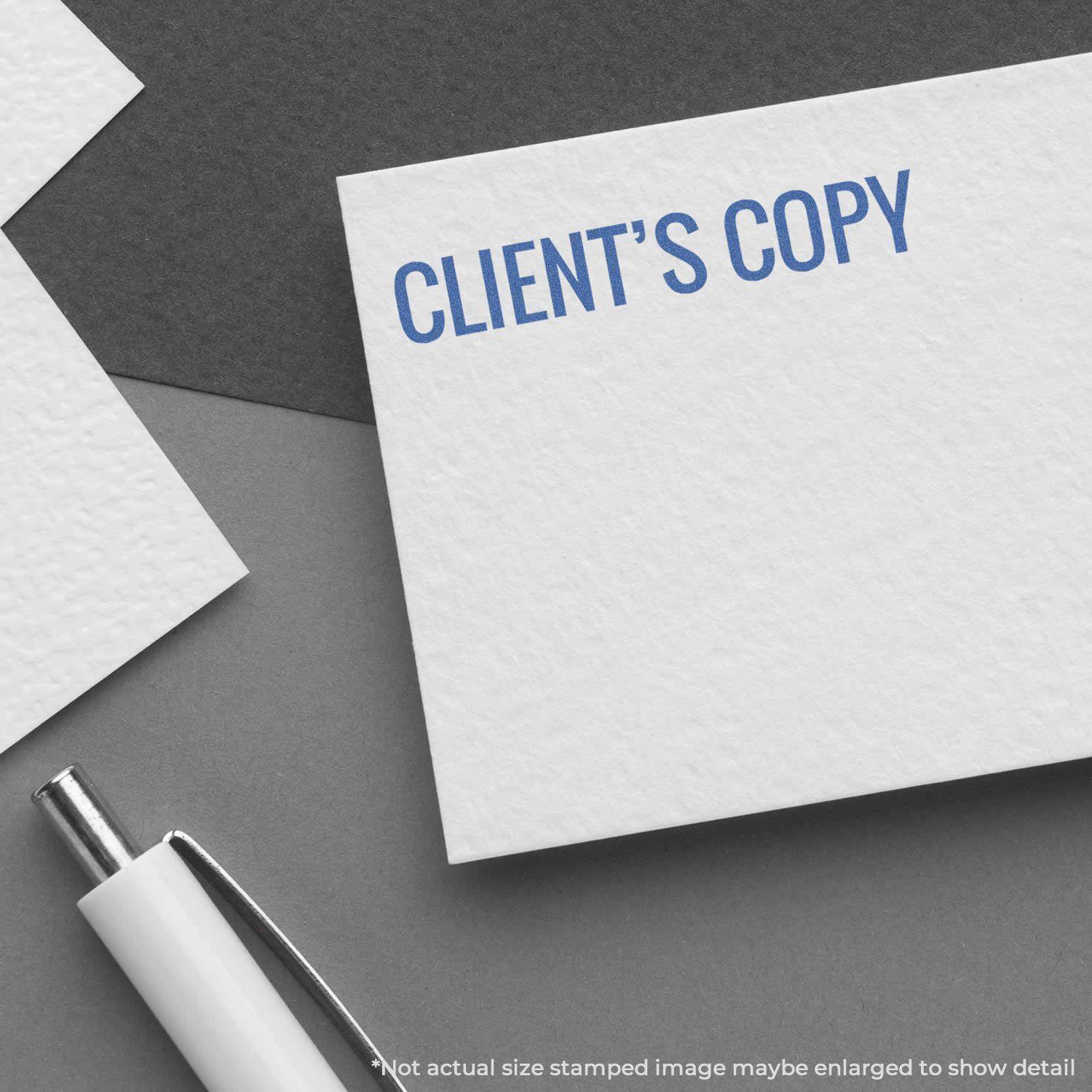 Client's Copy Xstamper Stamp in blue ink on white paper, with a white pen nearby on a dark gray surface.