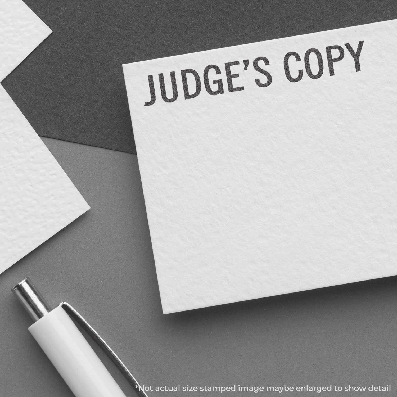 Judge's Copy Xstamper Stamp imprint on white paper, next to a pen, with text Not actual size stamped image maybe enlarged to show detail.