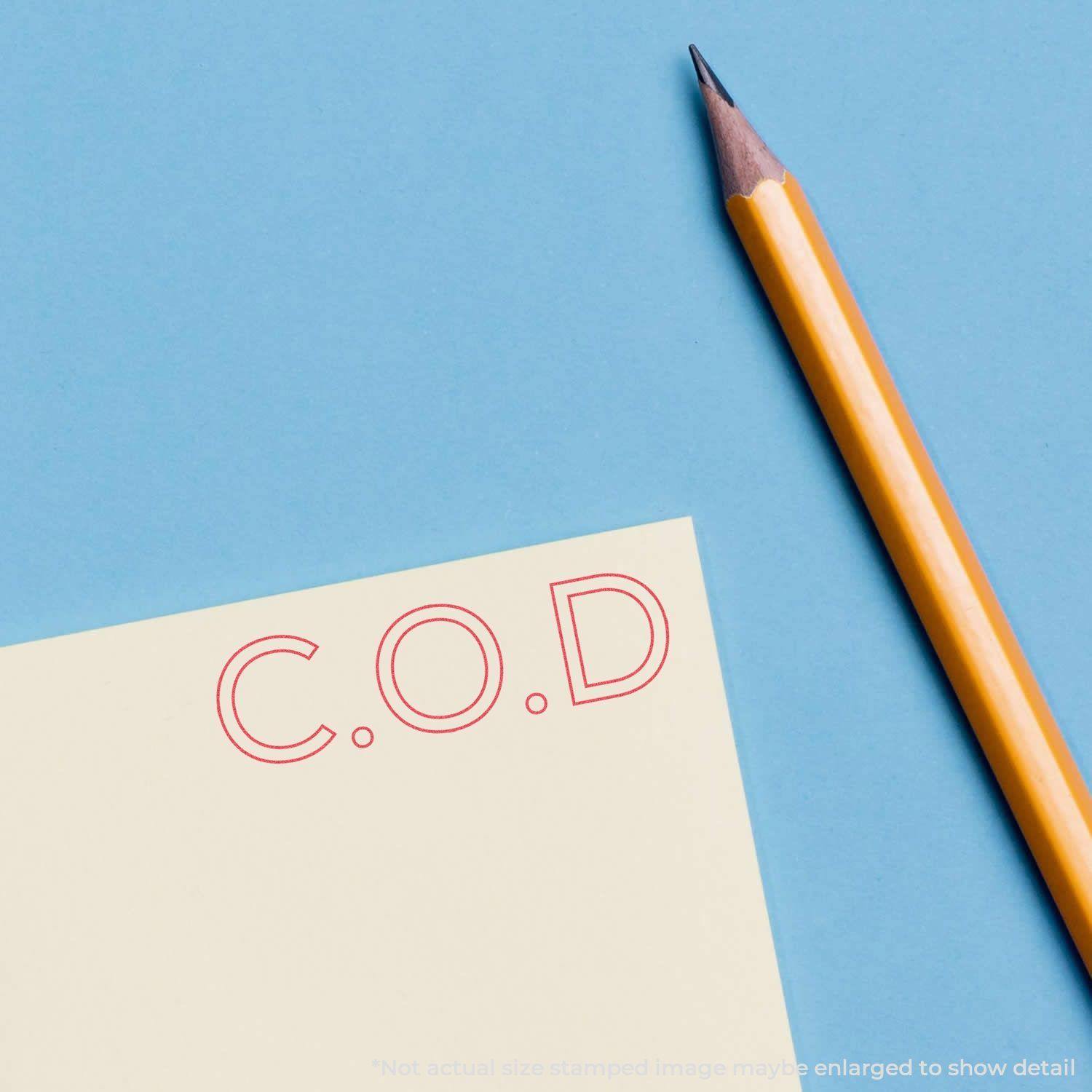 Red C.O.D. Xstamper Stamp imprint on a yellow paper next to a sharpened pencil on a blue background.