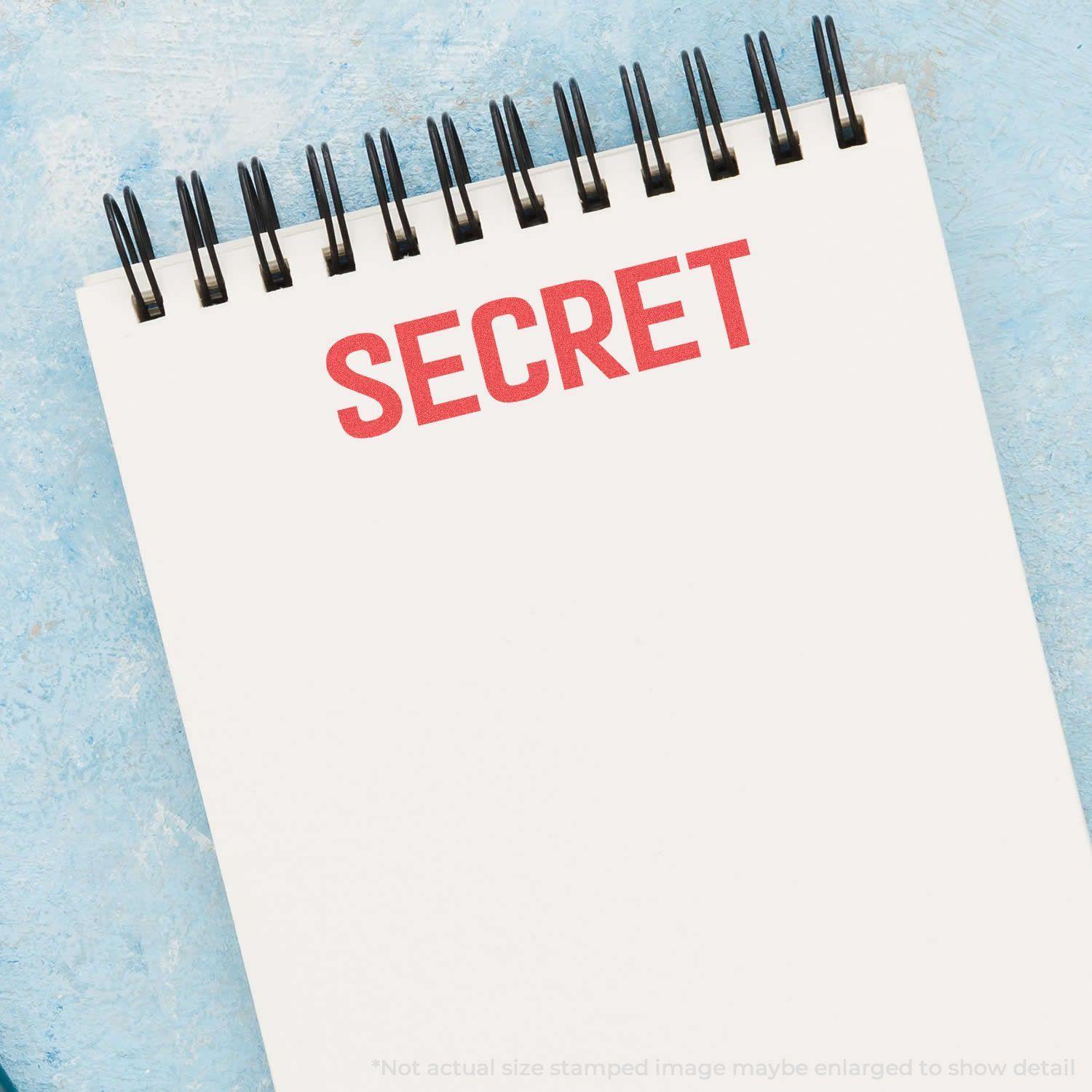 A notebook with SECRET stamped in red using the Secret Xstamper Stamp, placed on a light blue textured surface.