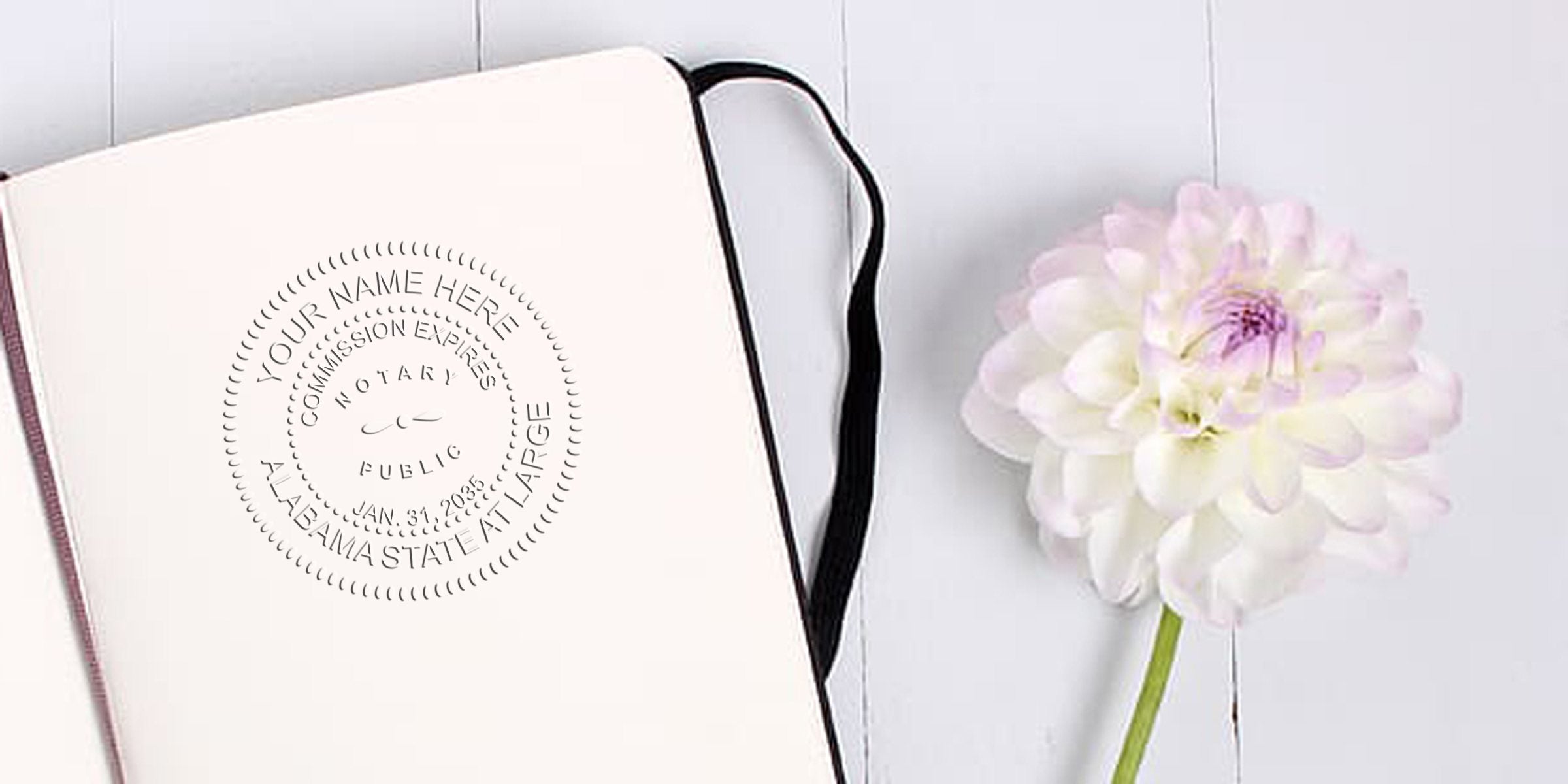 Open blank notebook with black elastic and embossed circular notary-style seal on the page beside a pale pink-white dahlia on white wooden background