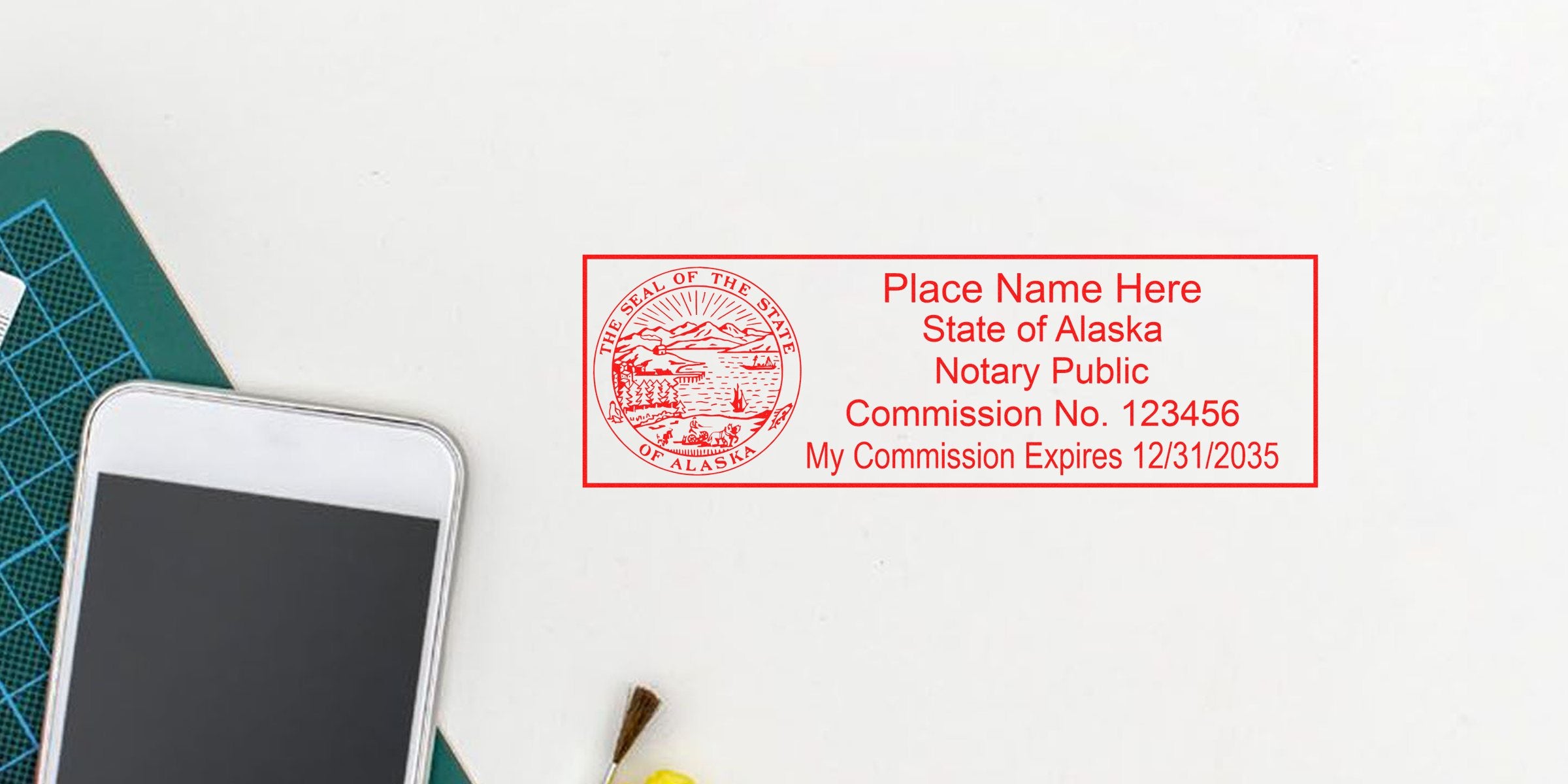Red rectangular Alaska notary public stamp mockup with state seal, placeholder name, commission number and expiration date on white background next to smartphone and cutting mat