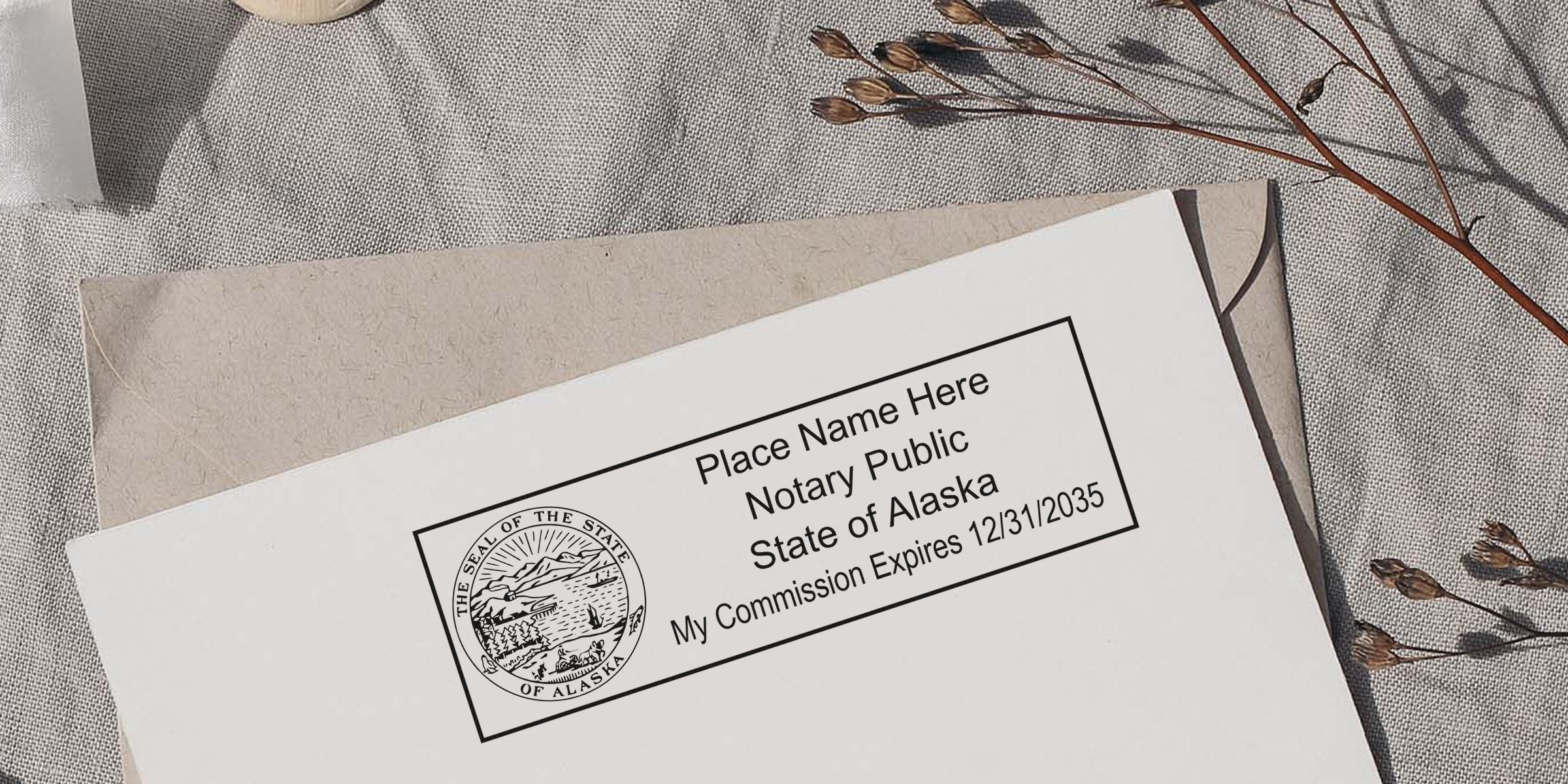 Alaska notary stamp sample on white paper showing state seal, "Notary Public" text and commission expiry, resting on a beige envelope over a linen background