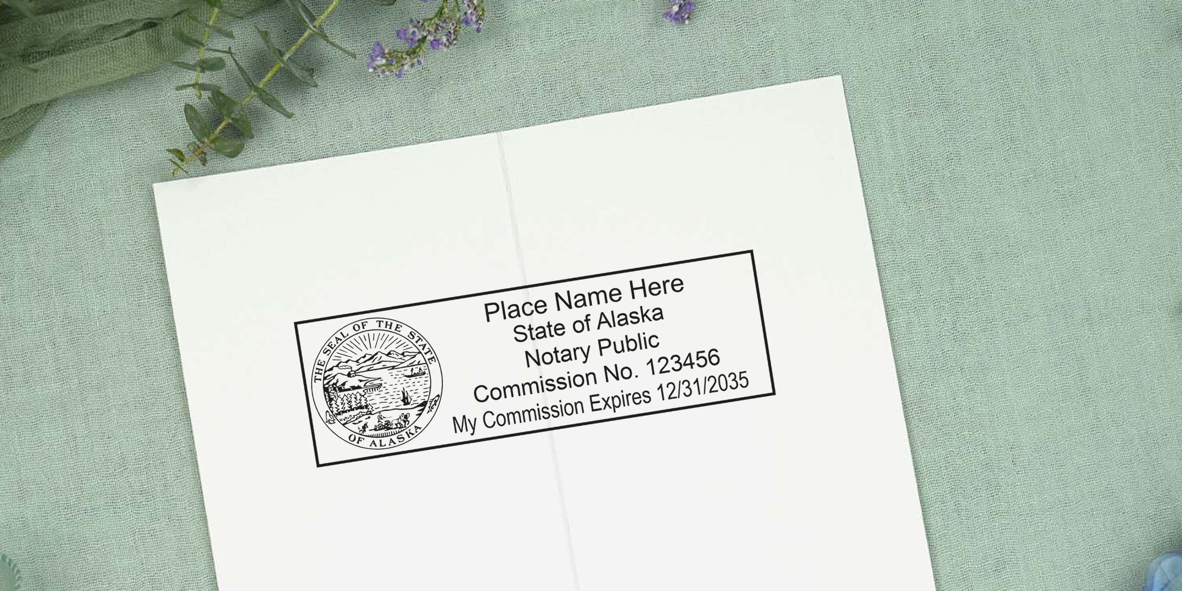 Alaska notary public stamp template on white paper showing state seal, placeholder name, commission no. 123456 and expiration 12/31/2035, photographed on green fabric with lavender accents