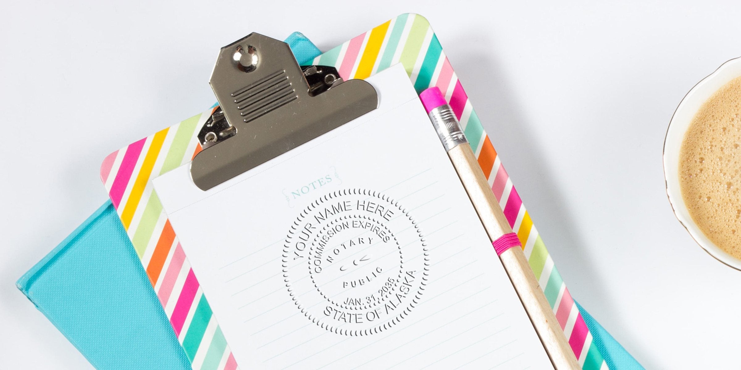 Top-down view of clipboard with colorful diagonal-striped edge holding a lined notepad stamped with a circular notary seal, wooden pencil with pink eraser, teal notebook beneath, and a cup of frothy coffee on a white background