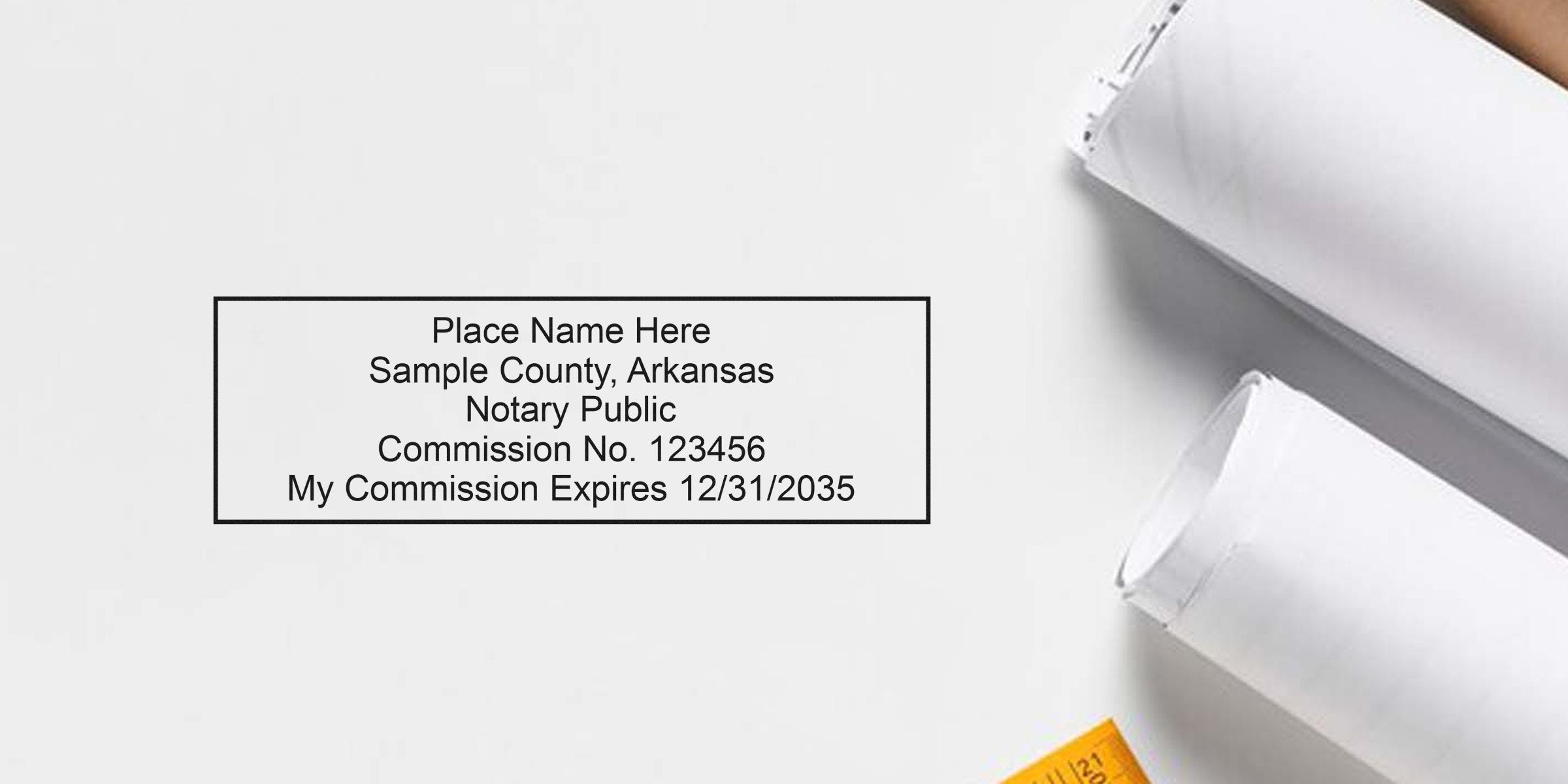 Rectangular notary stamp sample on white background reading "Place Name Here, Sample County, Arkansas, Notary Public, Commission No. 123456, My Commission Expires 12/31/2035" beside rolled architectural plans and a yellow measuring tape.