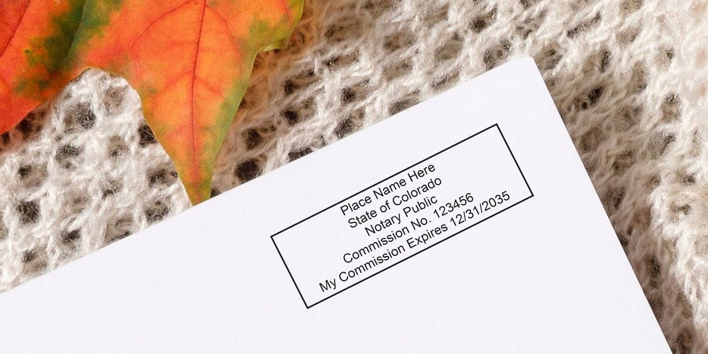 Close-up of a notary stamp on white paper reading "Place Name Here, State of Colorado, Notary Public, Commission No. 123456, My Commission Expires 12/31/2035" resting on a cream knit blanket with an orange autumn leaf.