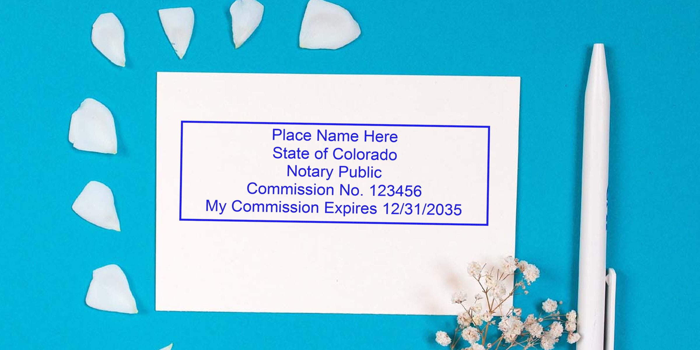 White notary stamp sample card reading "Place Name Here State of Colorado Notary Public Commission No. 123456 My Commission Expires 12/31/2035" on turquoise background with white pen, scattered petals and baby's breath flowers.