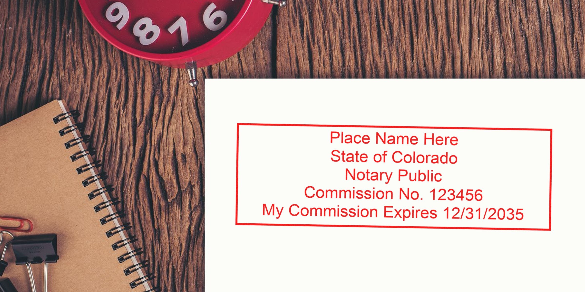 Overhead photo of a rustic wood desk with a red alarm clock, spiral notebook and a paper displaying a red Colorado notary public stamp template with commission number and expiration date.