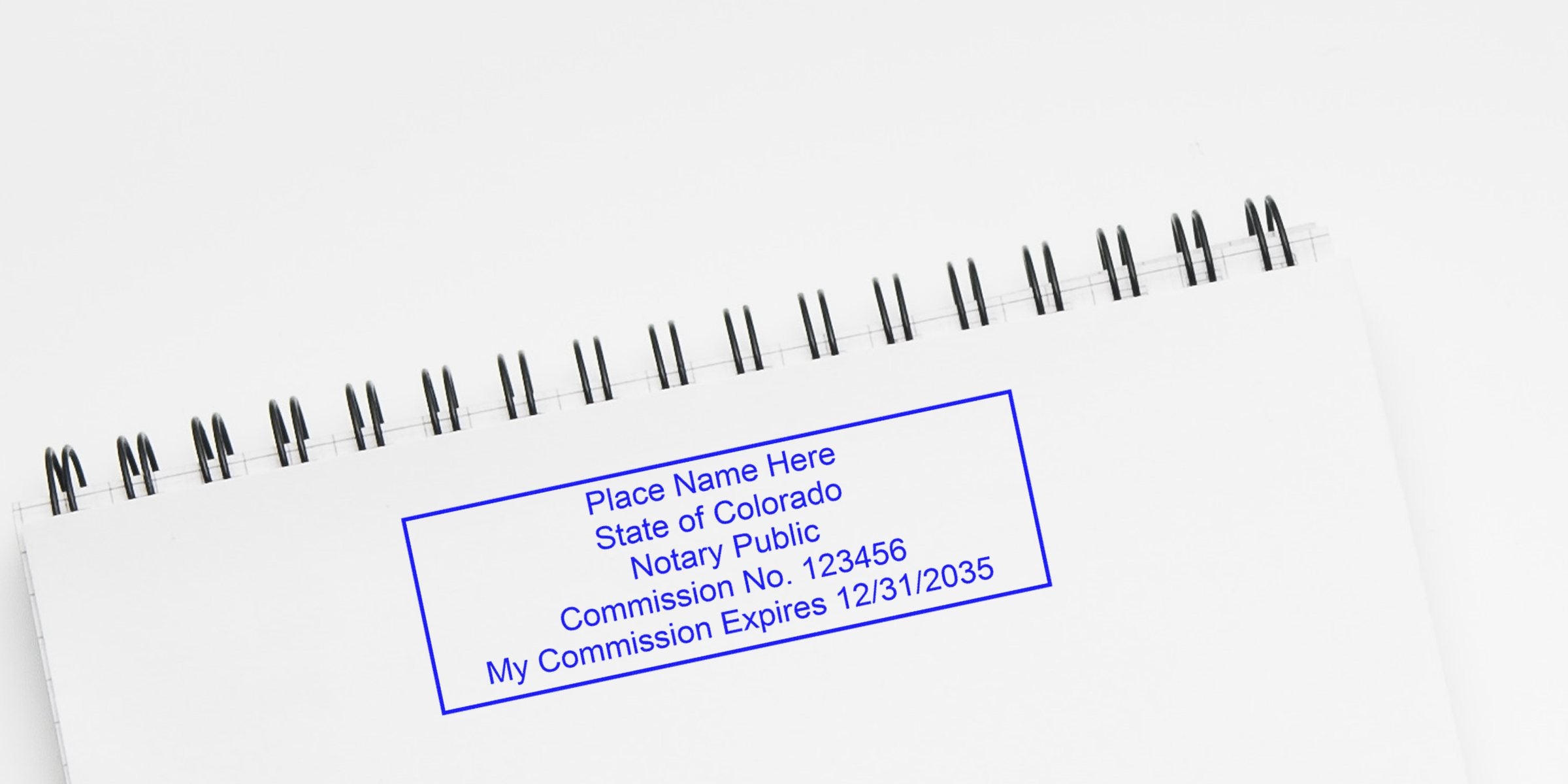Blue rectangular notary stamp on a white spiral-bound notebook reading "Place Name Here, State of Colorado, Notary Public, Commission No. 123456, My Commission Expires 12/31/2035" in a minimal flatlay background