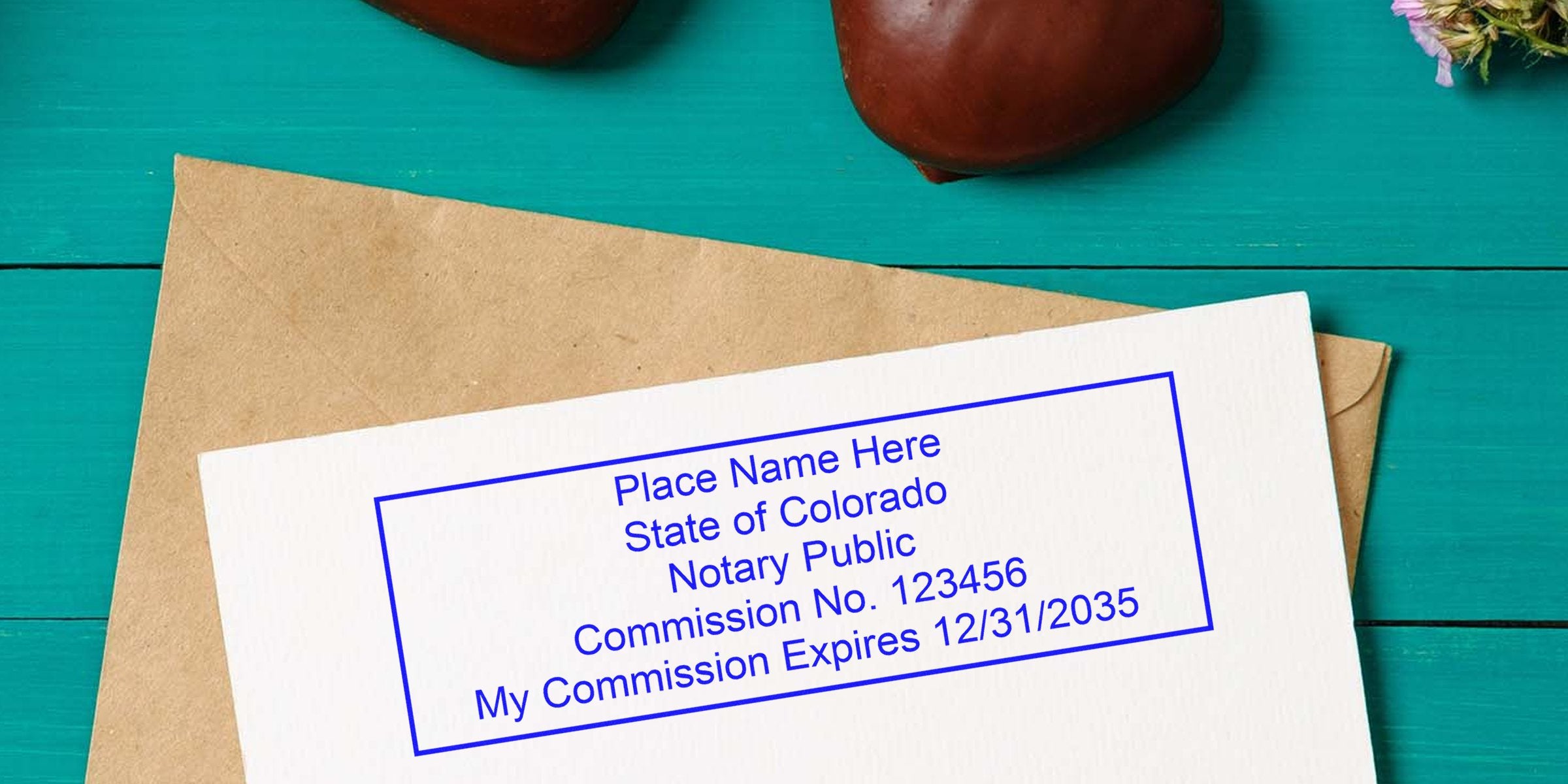 Notary stamp example on white card over kraft envelope on teal wooden table: blue rectangular Colorado Notary Public stamp showing commission number and expiration date.
