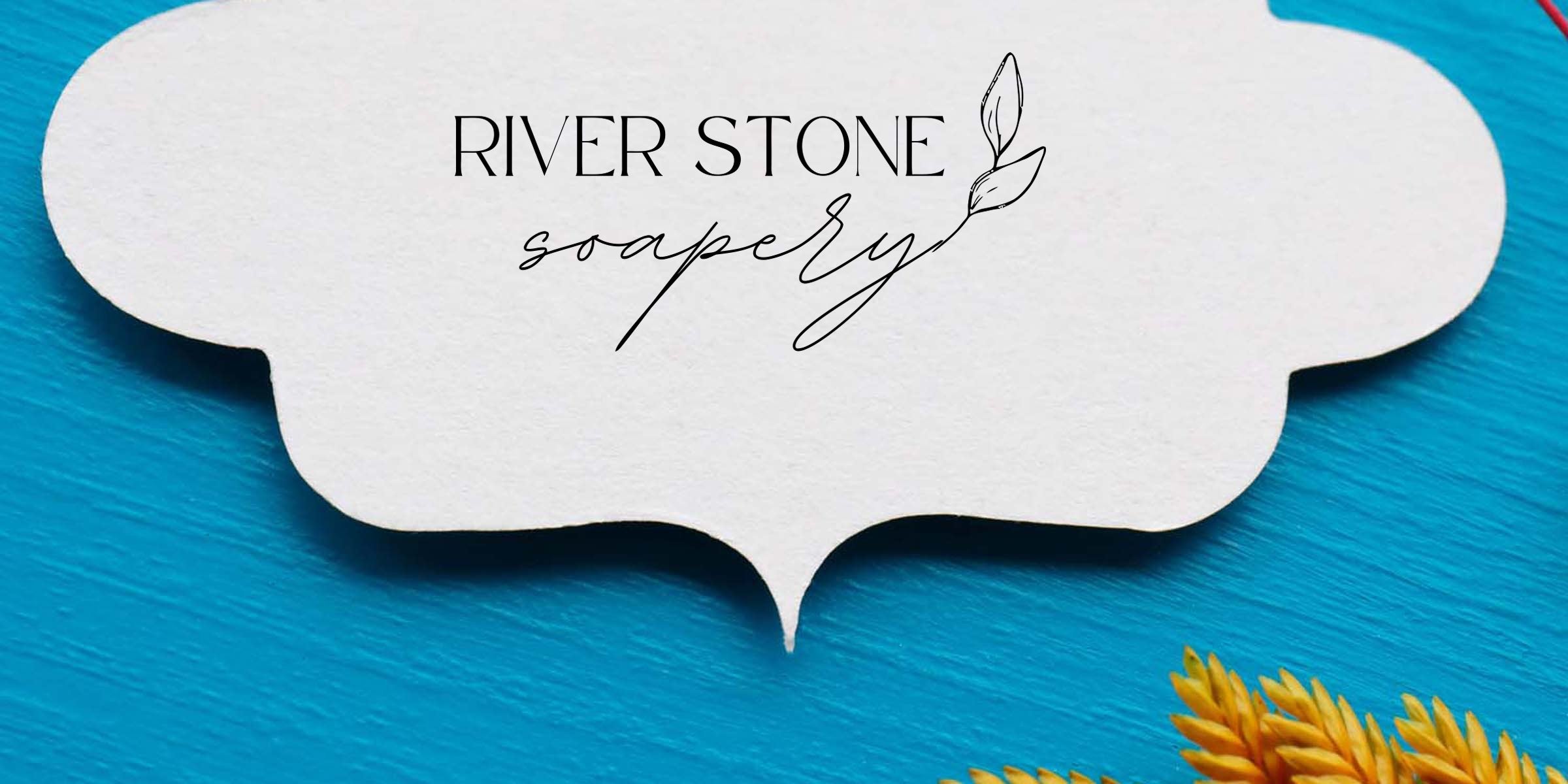 River Stone Soapery logo in elegant black script on a white decorative label over bright blue wooden background with golden wheat sprigs