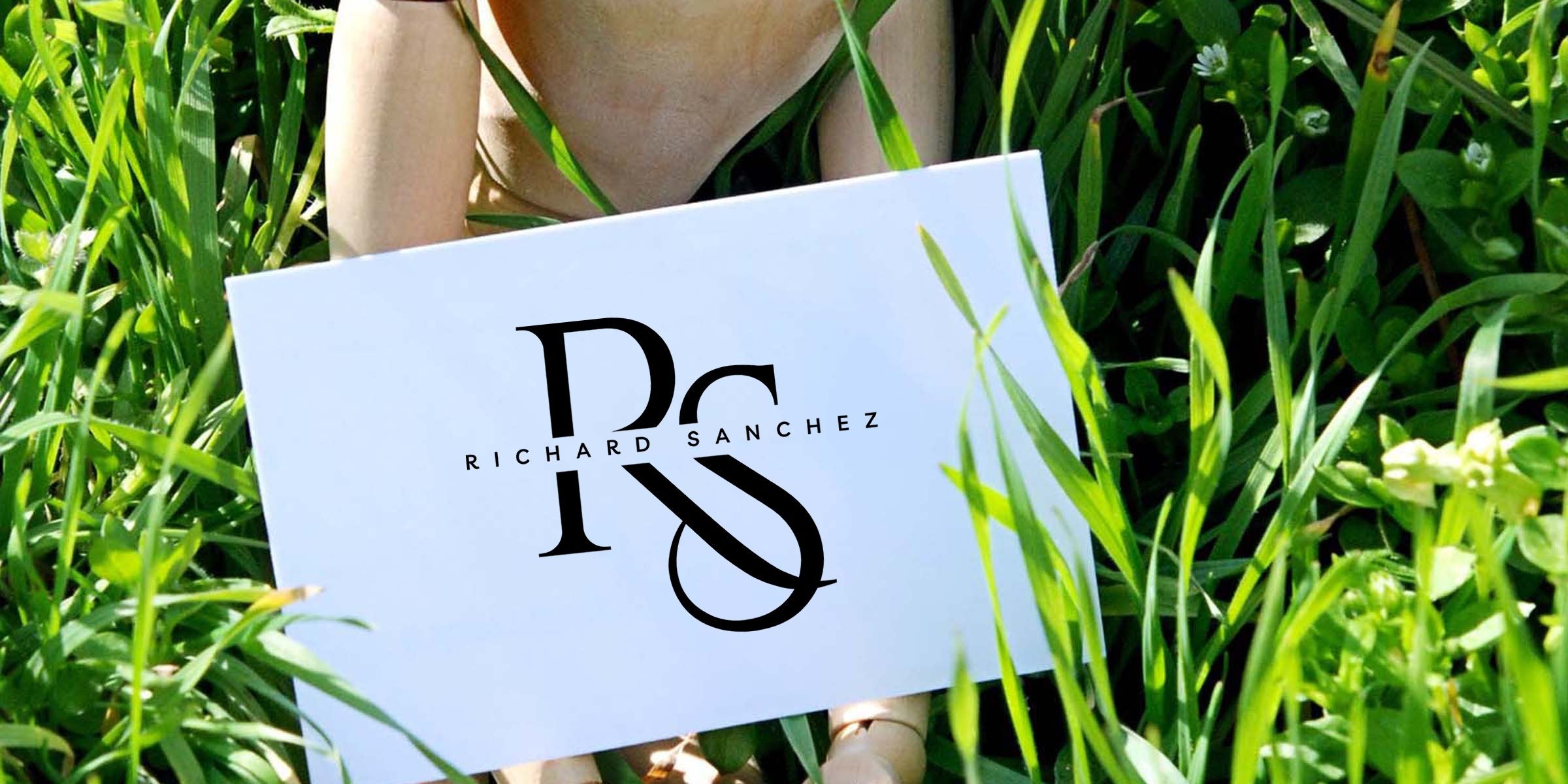 Person holding white card in tall green grass featuring a black stylized "RS" monogram and horizontal name text, minimalist branding mockup on a natural outdoor background.