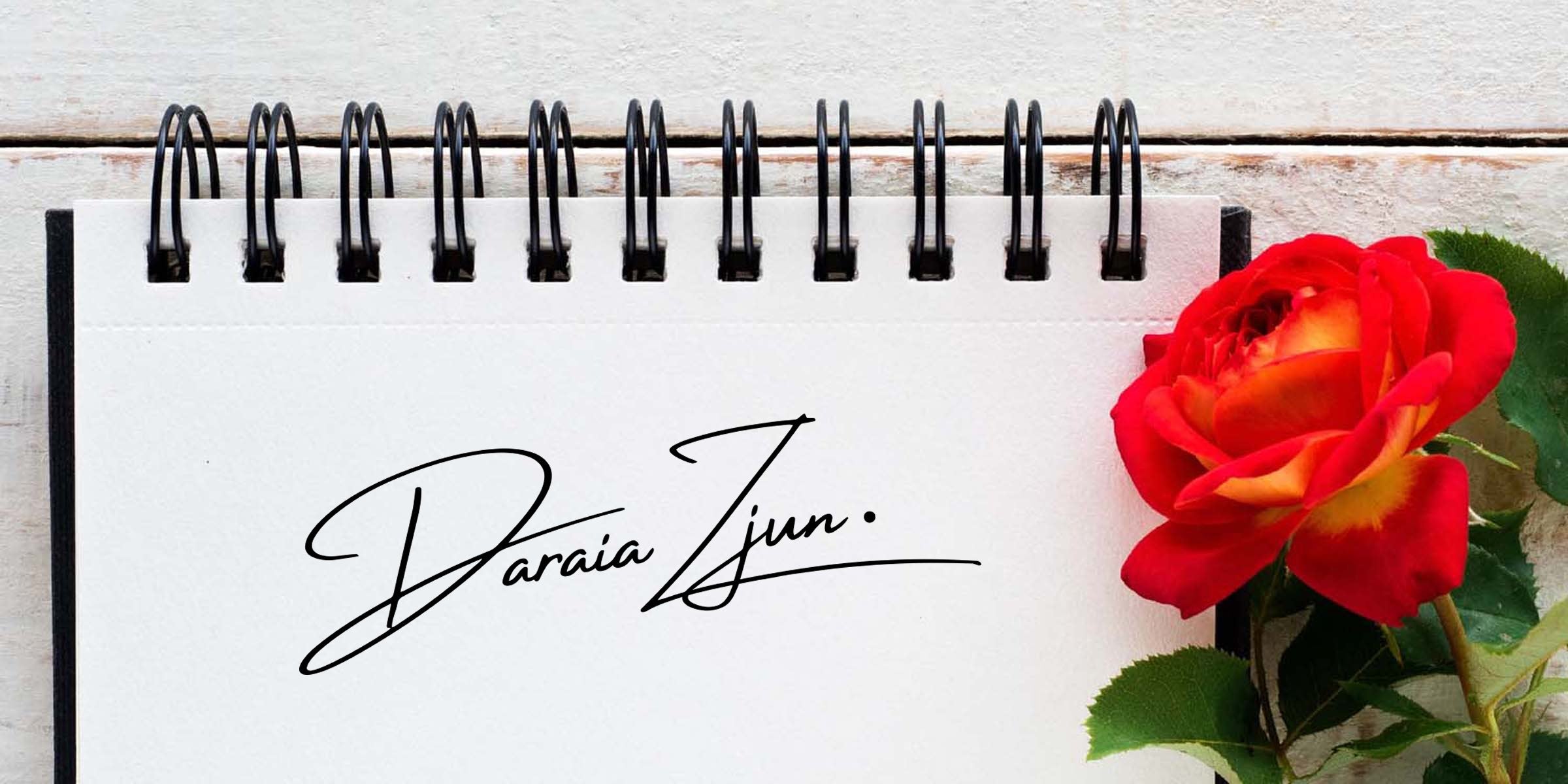 Spiral-bound white notepad with black handwritten signature on the page, red-yellow rose beside it on a white wooden background — signature mockup for branding and stationery design