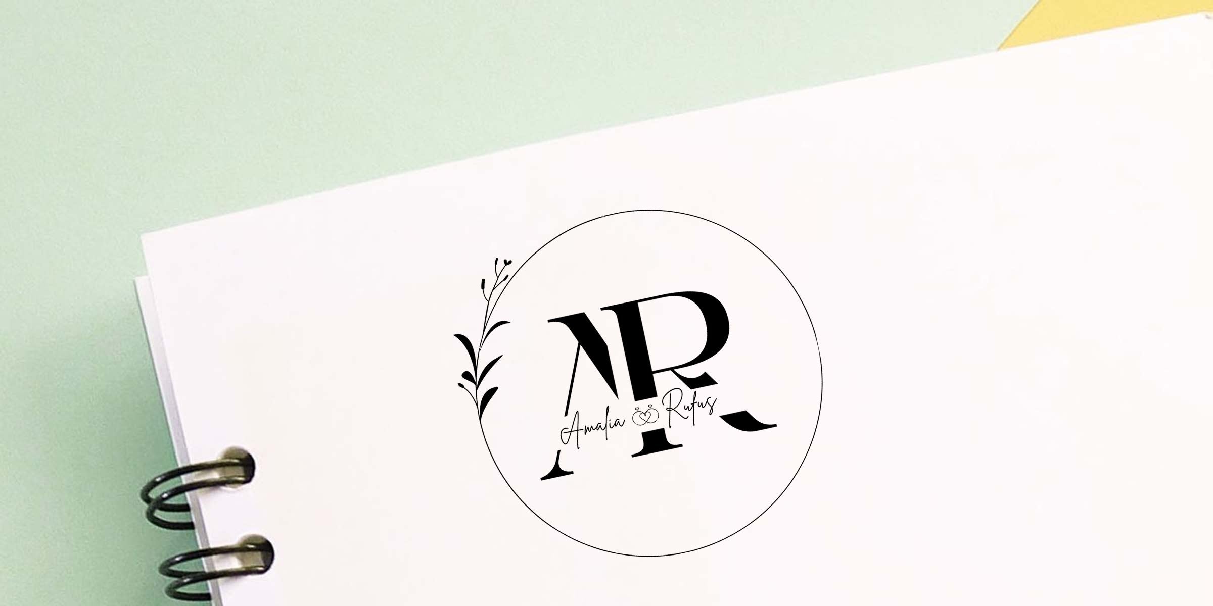 Minimalist black "AR" wedding monogram with floral accent and interlocked rings on white notebook cover, elegant circular logo mockup for Amalia & Rufus against a soft pastel green background