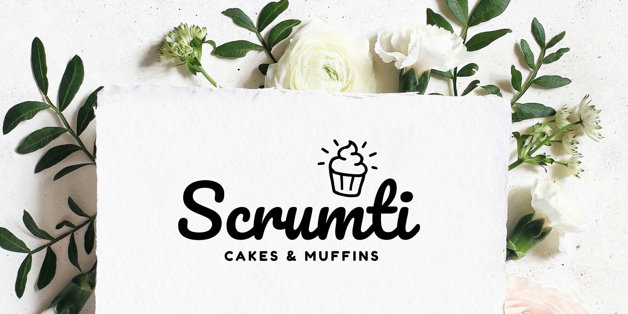 Scrumti Cakes & Muffins black logo with cupcake icon on white textured paper, styled flatlay with green leaves and white flowers for bakery branding