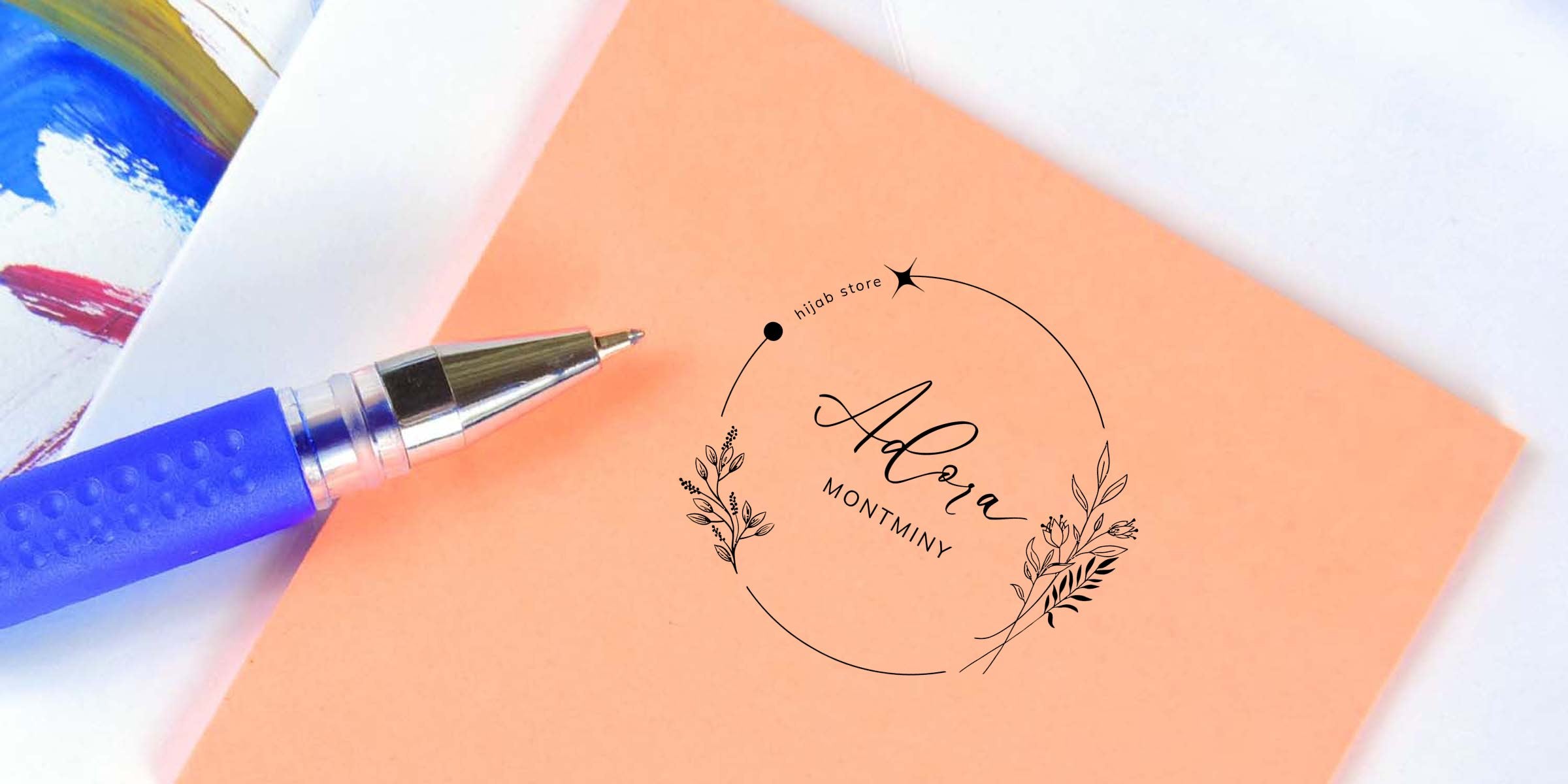 Close-up of a blue gel pen resting on a peach notecard featuring a circular floral logo reading "Adora Montminy" with small "hijab store" text and colorful paint swatches at the edge.