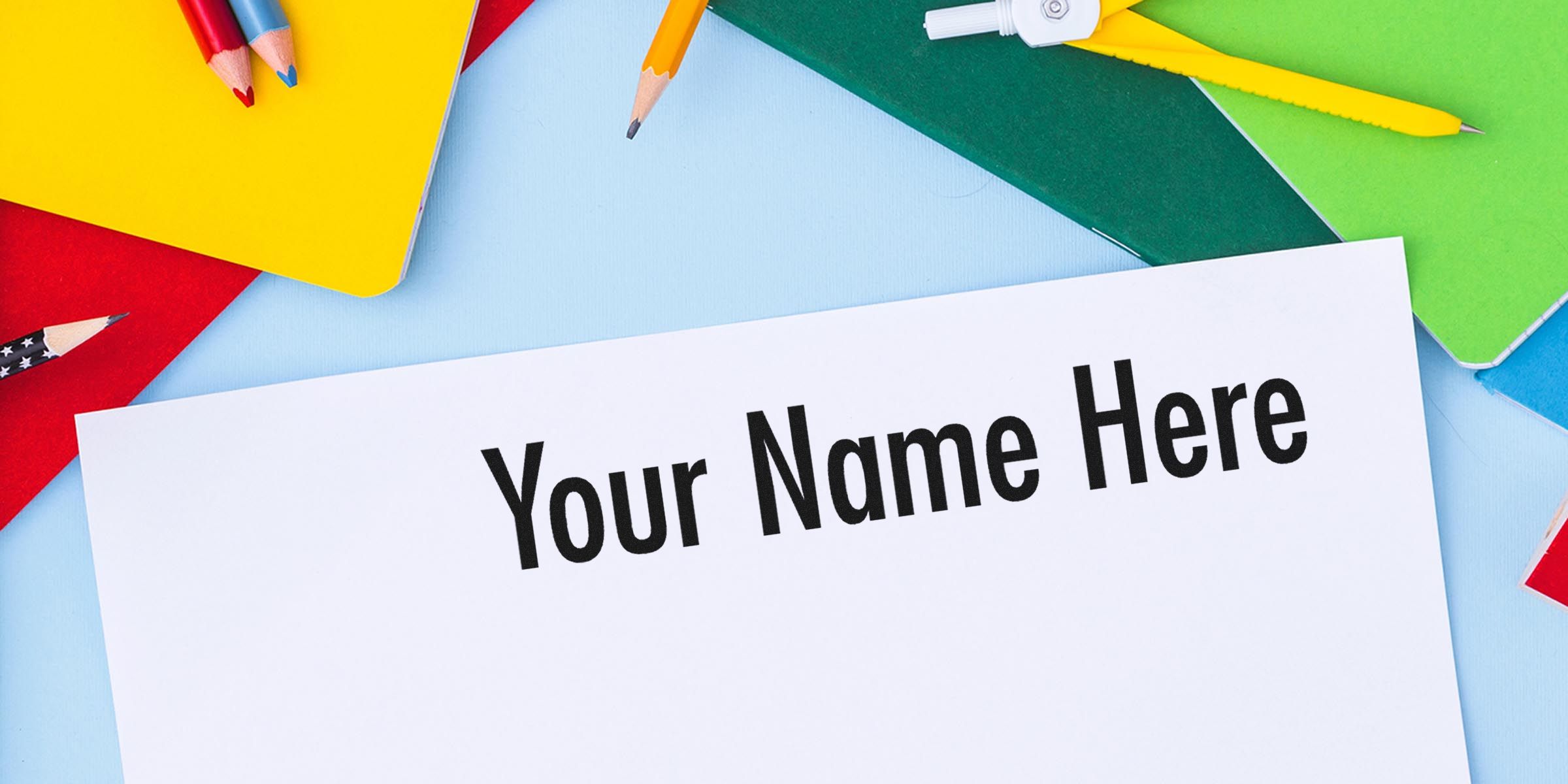 A white sheet with the words “Your Name Here” in bold black letters, surrounded by colorful notebooks, pencils, and other school supplies.