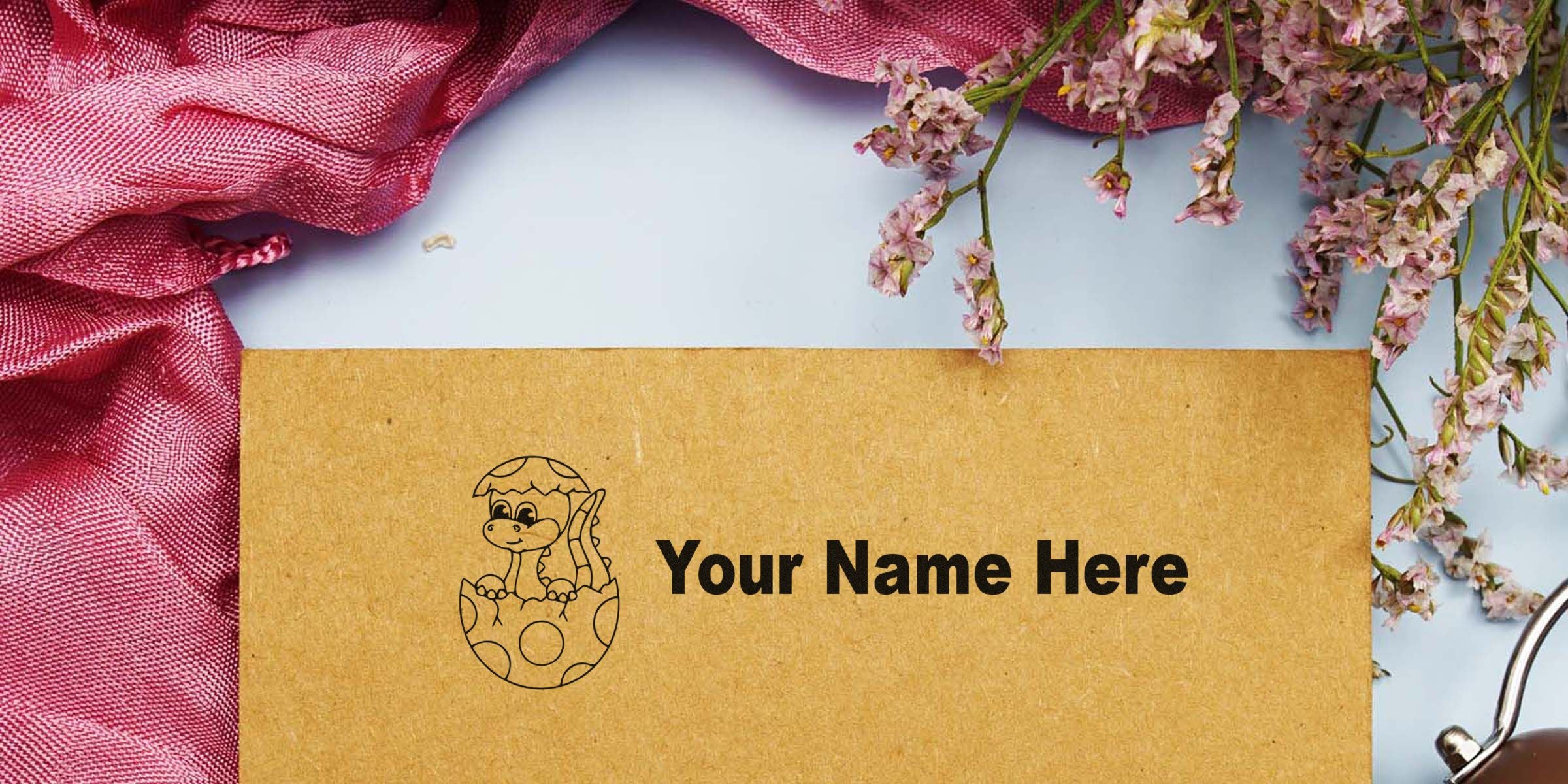 Brown kraft card reading “Your Name Here” with a small dinosaur illustration emerging from a spotted egg, set against pink fabric and flower accents.
