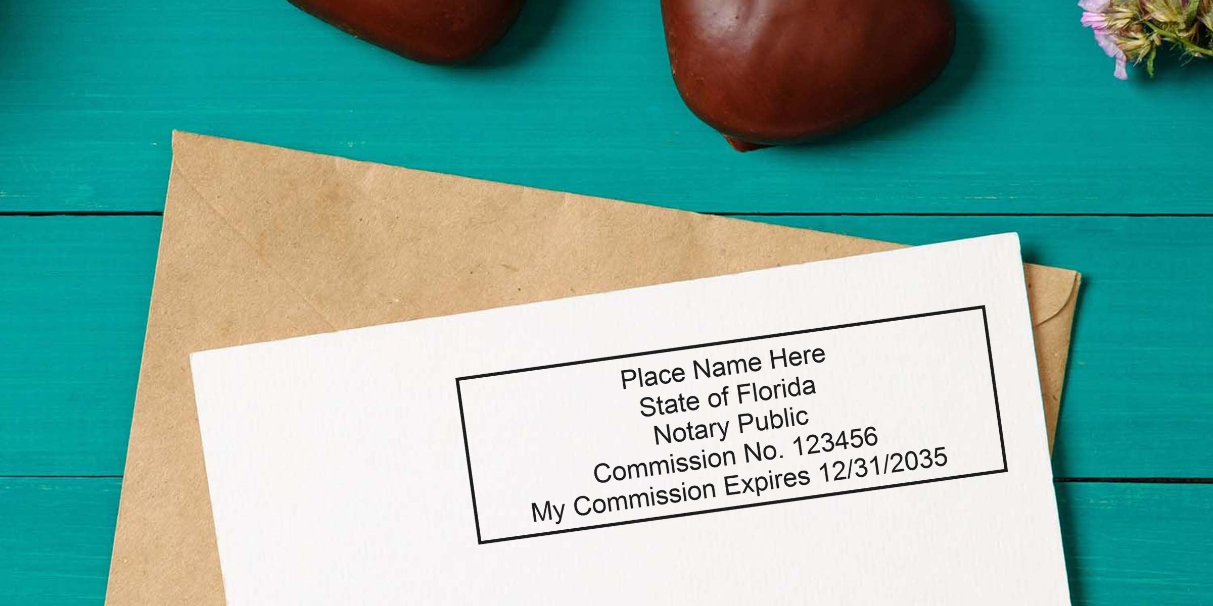 Close-up of a Florida notary public stamp sample on white paper showing commission number and expiration date, on a kraft envelope over teal wooden table, decorative chestnuts and flowers.