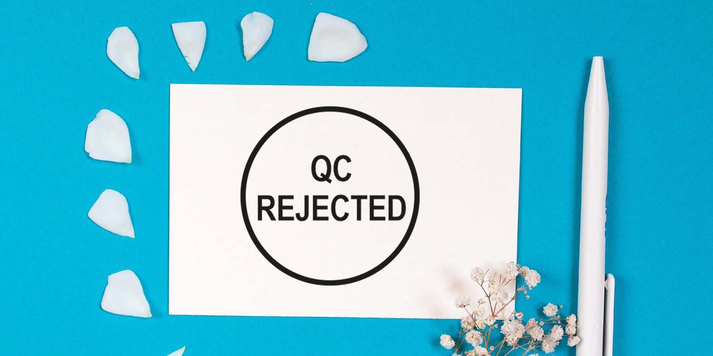Alt text: A white card on a blue background with a black circle containing the words “QC REJECTED”; white petals and a white pen decor surround the card.