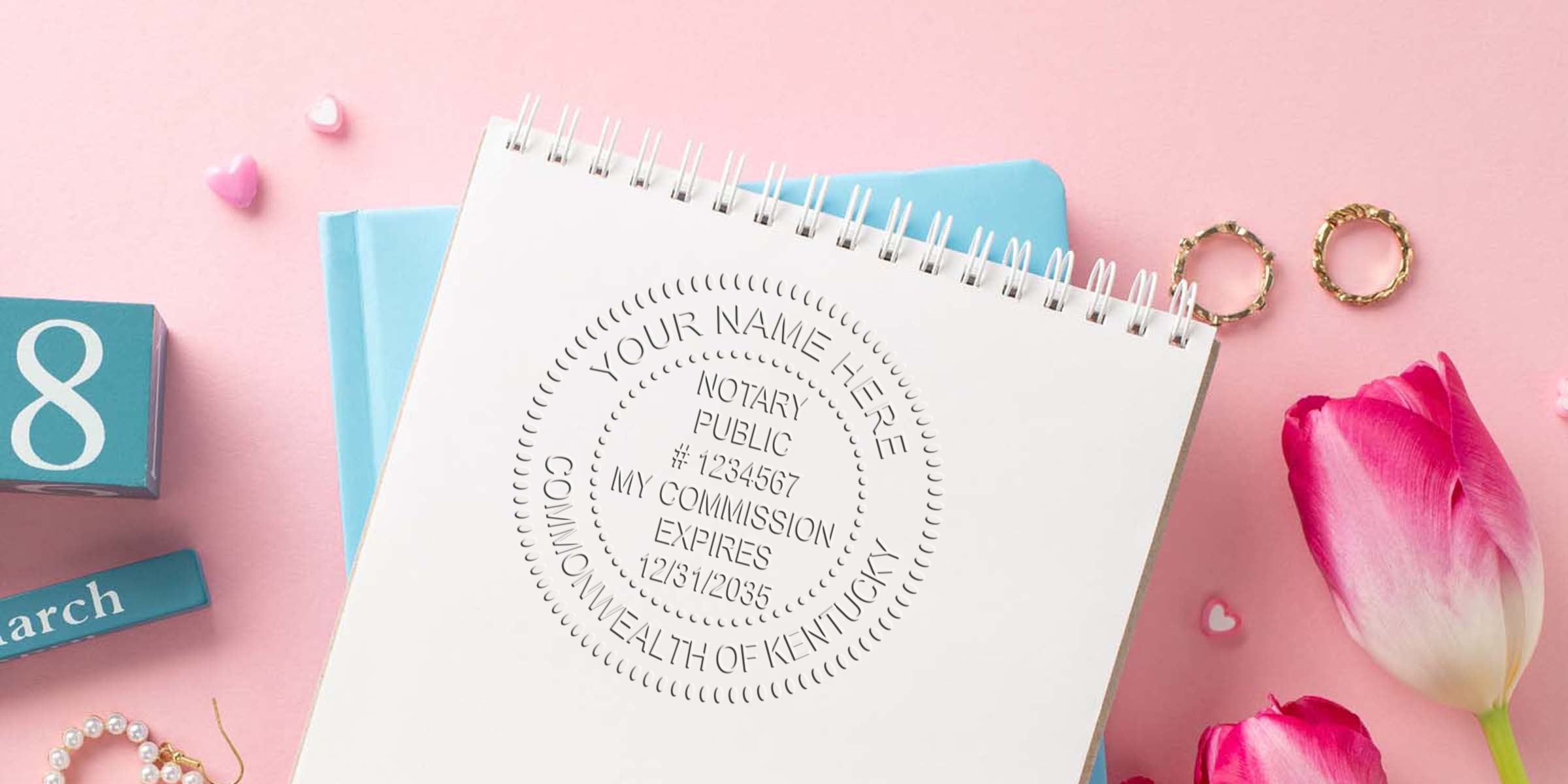 Flat lay of spiral notebook with embossed notary public seal mockup, pink tulips, gold rings, pearl bracelet and blue March calendar block on pastel pink background — feminine business stationery flat lay