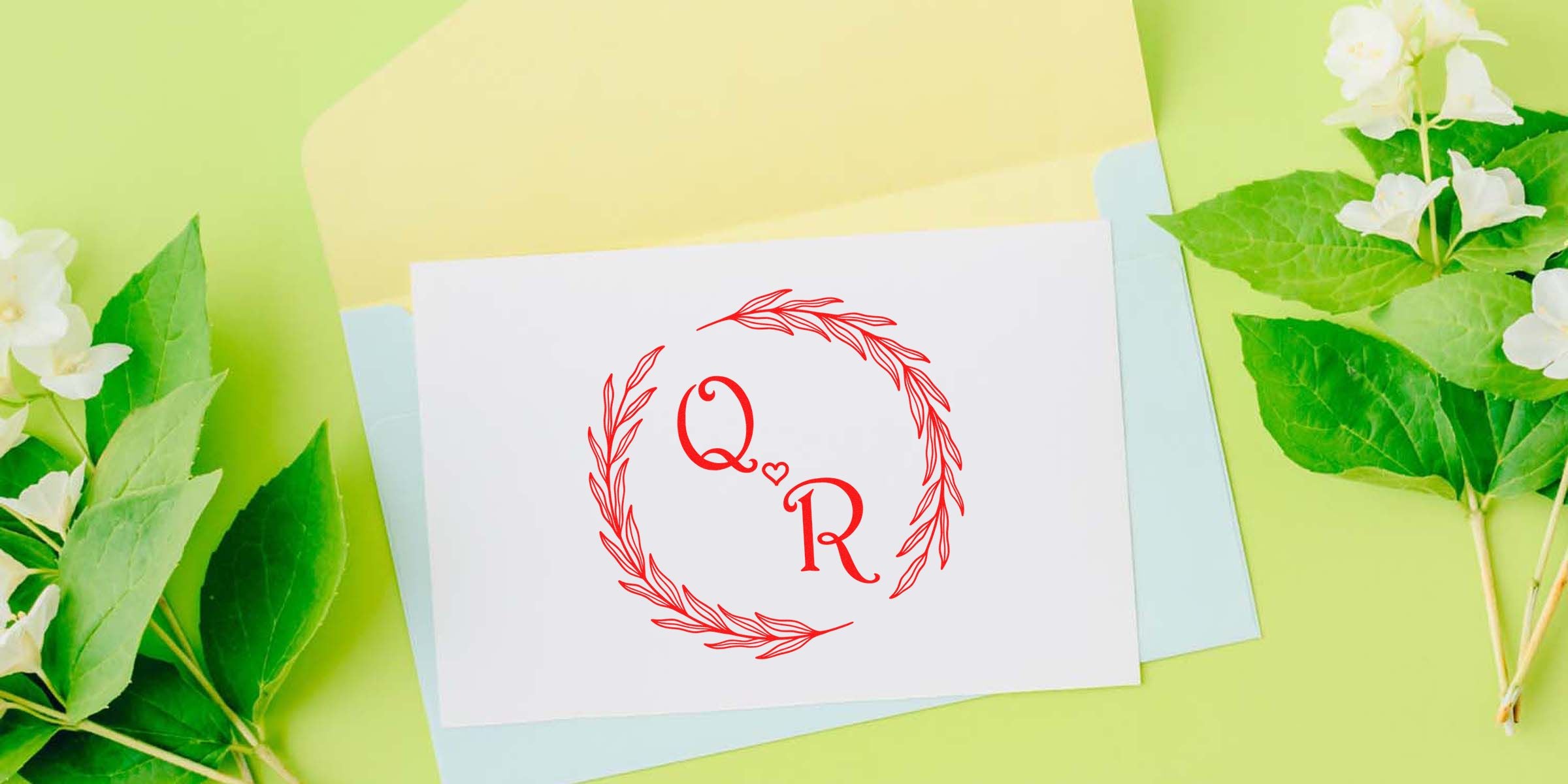 White card featuring a red Q and R monogram with a small heart between them, encircled by a red laurel wreath, placed on pastel papers with green leaves and white flowers in the background.