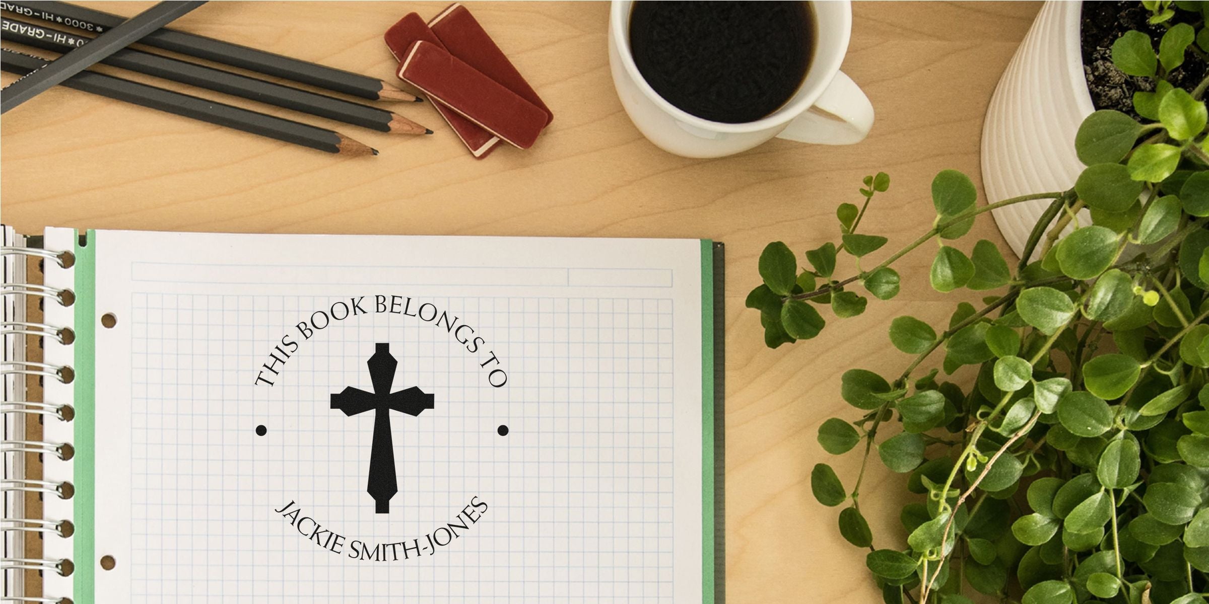 Open notebook with a circular logo reading “This Book Belongs To Jackie Smith-Jones” around a cross, on a wooden desk with pencils, a mug of coffee, and green plants nearby.