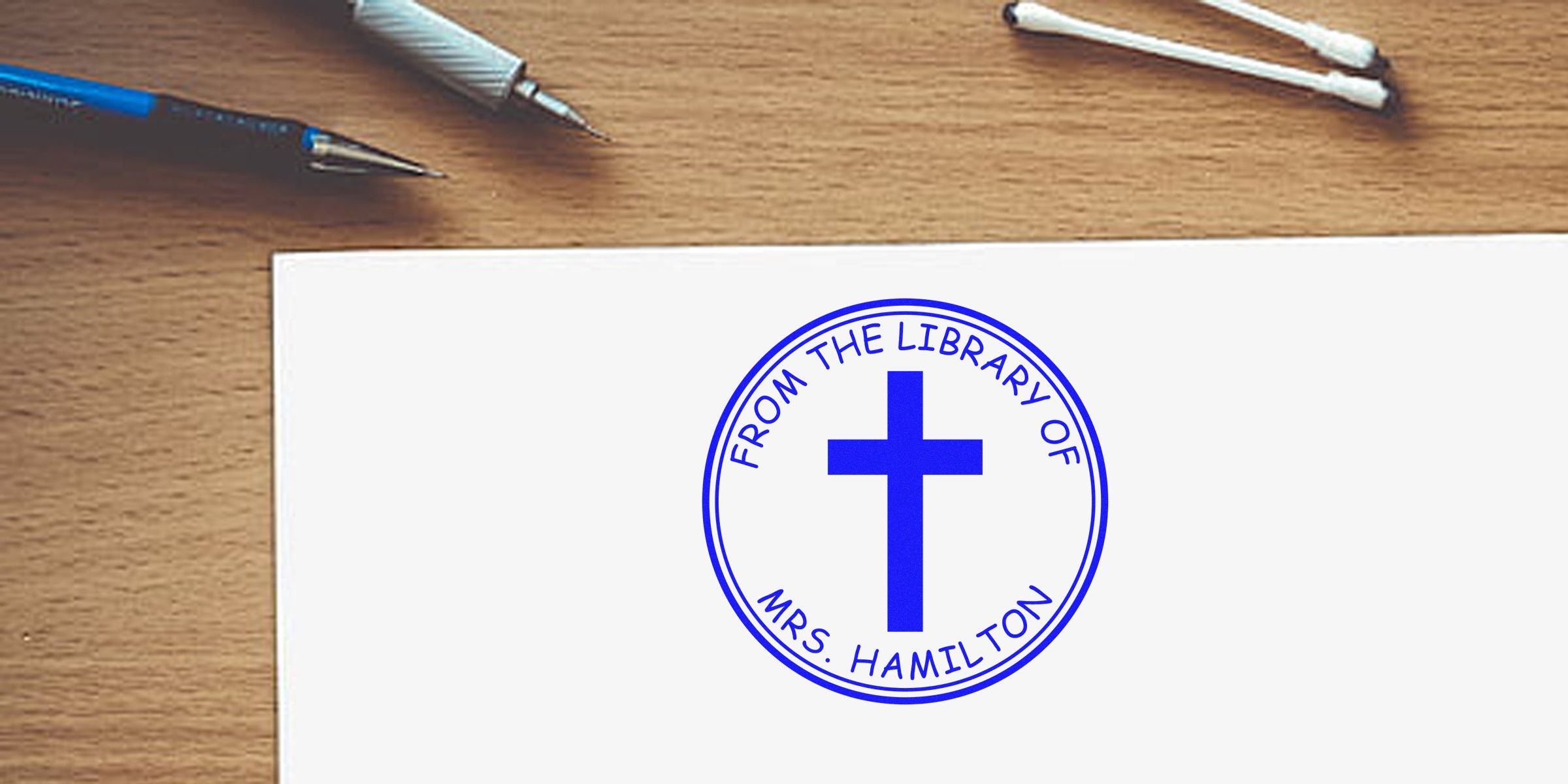 Blue circular stamp reading "From the Library of Mrs. Hamilton" with a cross in the center on a white sheet, placed on a wooden desk with pens nearby.